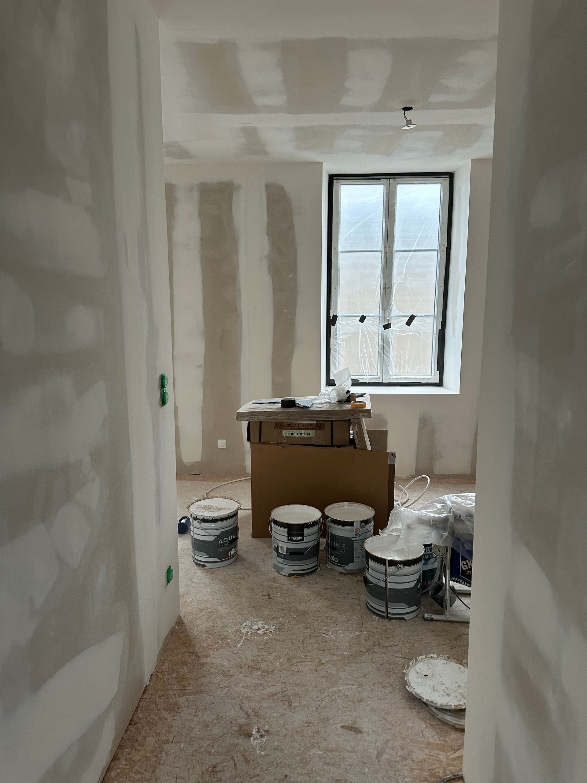 Pièce inachevée avec des plaques de plâtre prêtes à peindre, des seaux de peinture et une fenêtre.