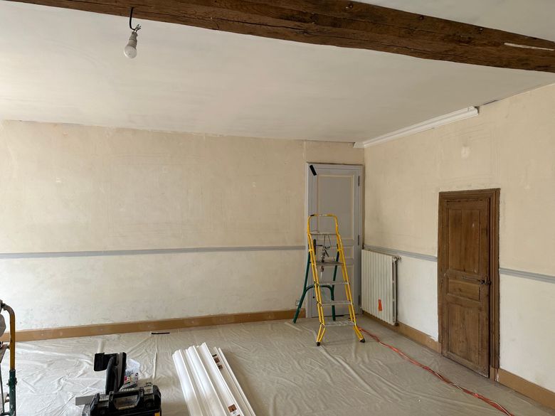 Pièce vide en cours de rénovation avec échelle, matériel de peinture, poutres apparentes.