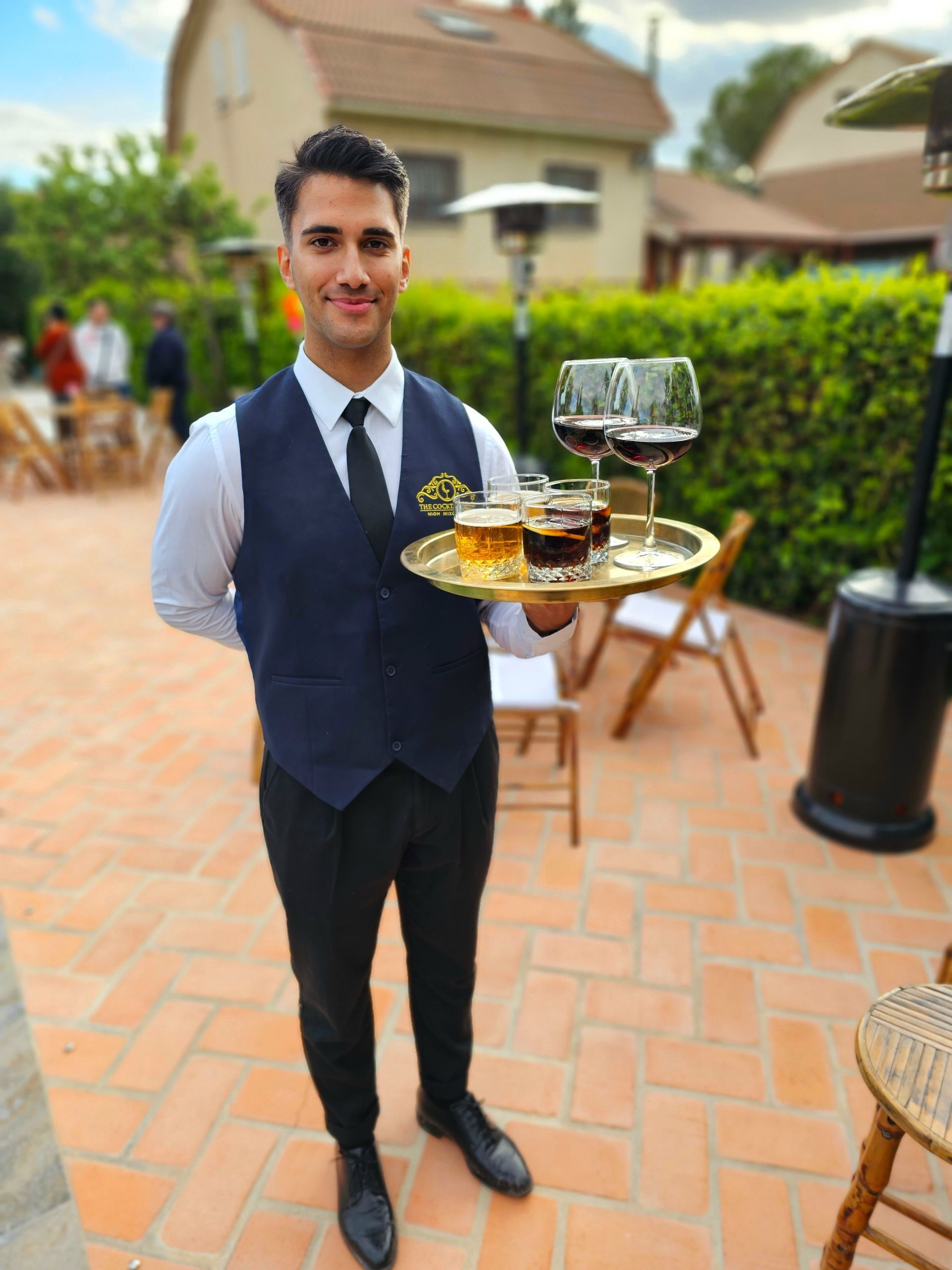 El camarero sostiene una bandeja de bebidas al aire libre, vestido con chaleco y corbata.