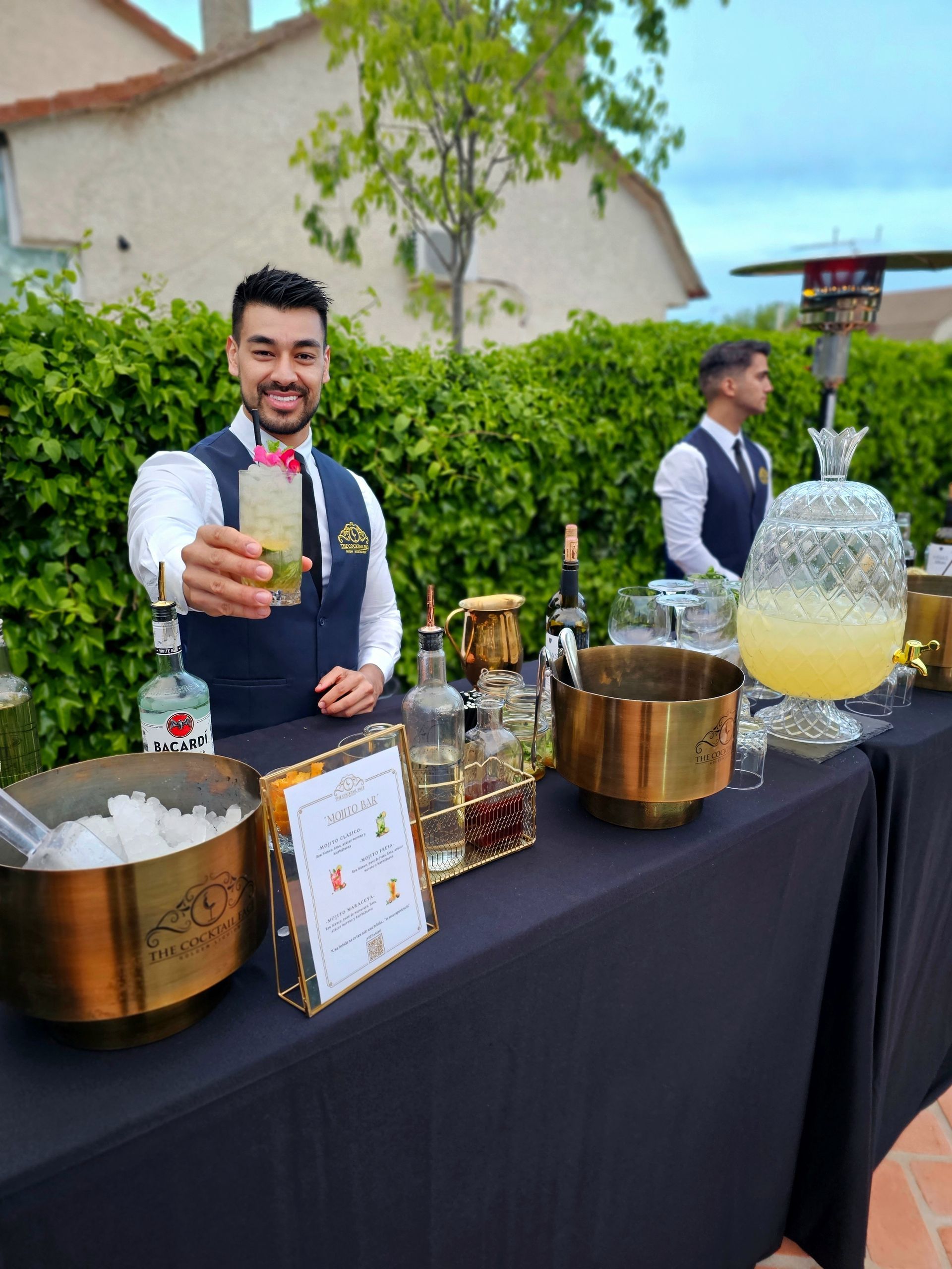 Camarero sirviendo un cóctel en un bar al aire libre con detalles dorados.