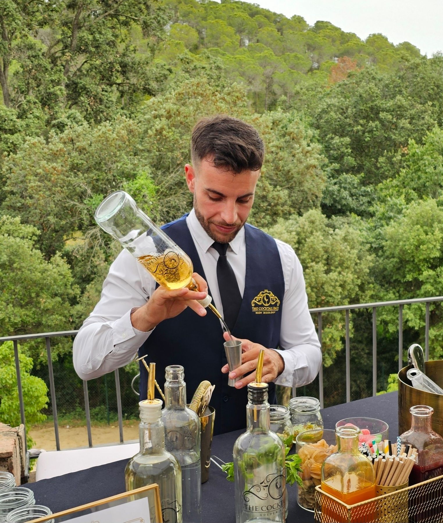 Un camarero sirve una copa, vestido con chaleco y corbata, al aire libre con un bosque de fondo.