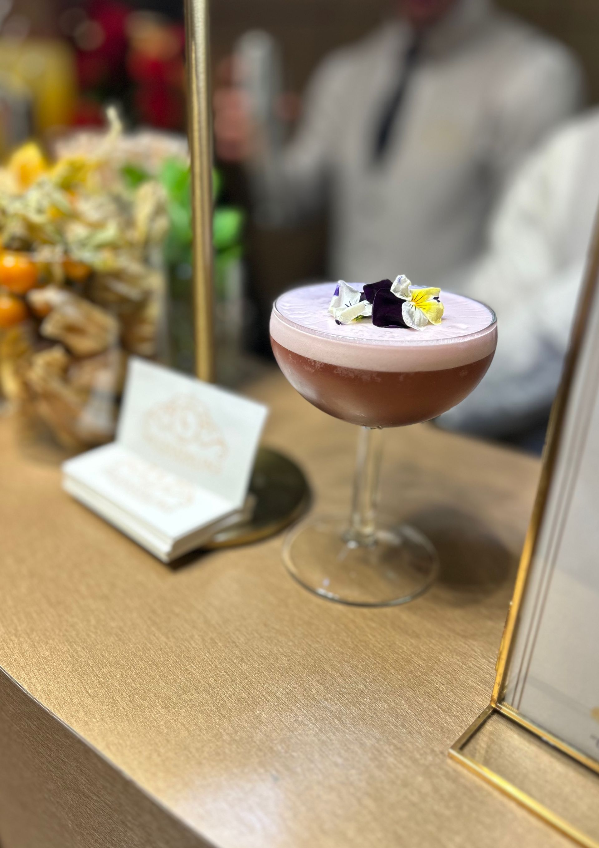 Cóctel en copa de tallo con decoración floral sobre barra dorada.