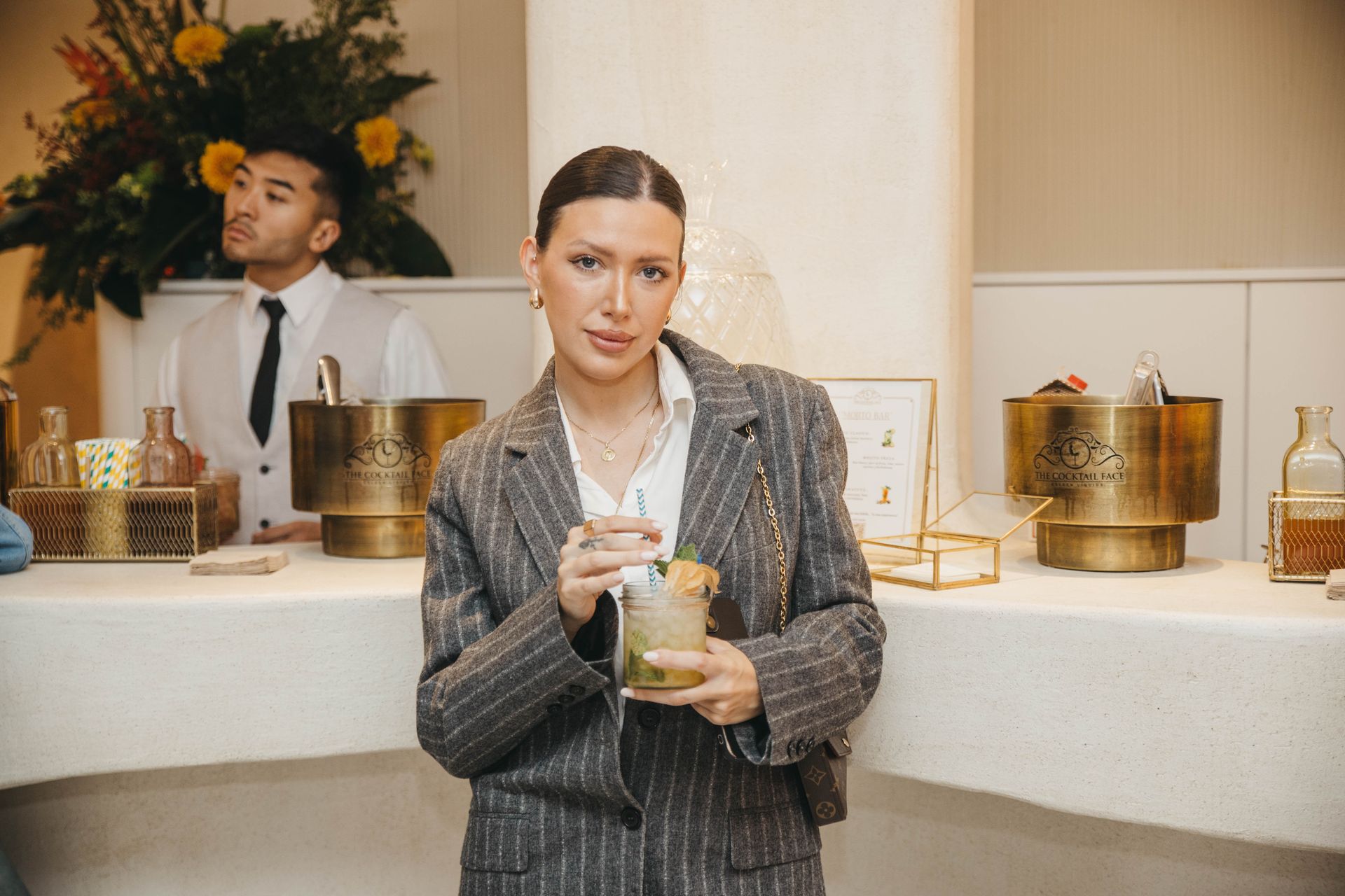Mujer con traje gris sostiene una bebida en un bar; camarero detrás de ella.