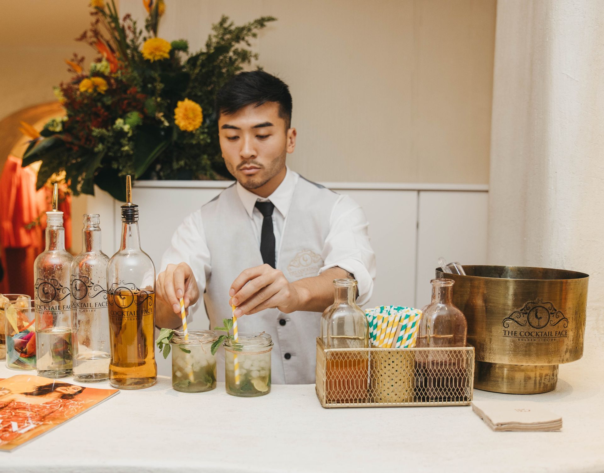 Un camarero prepara bebidas en un bar, removiendo con una pajita. Se ven botellas y flores.