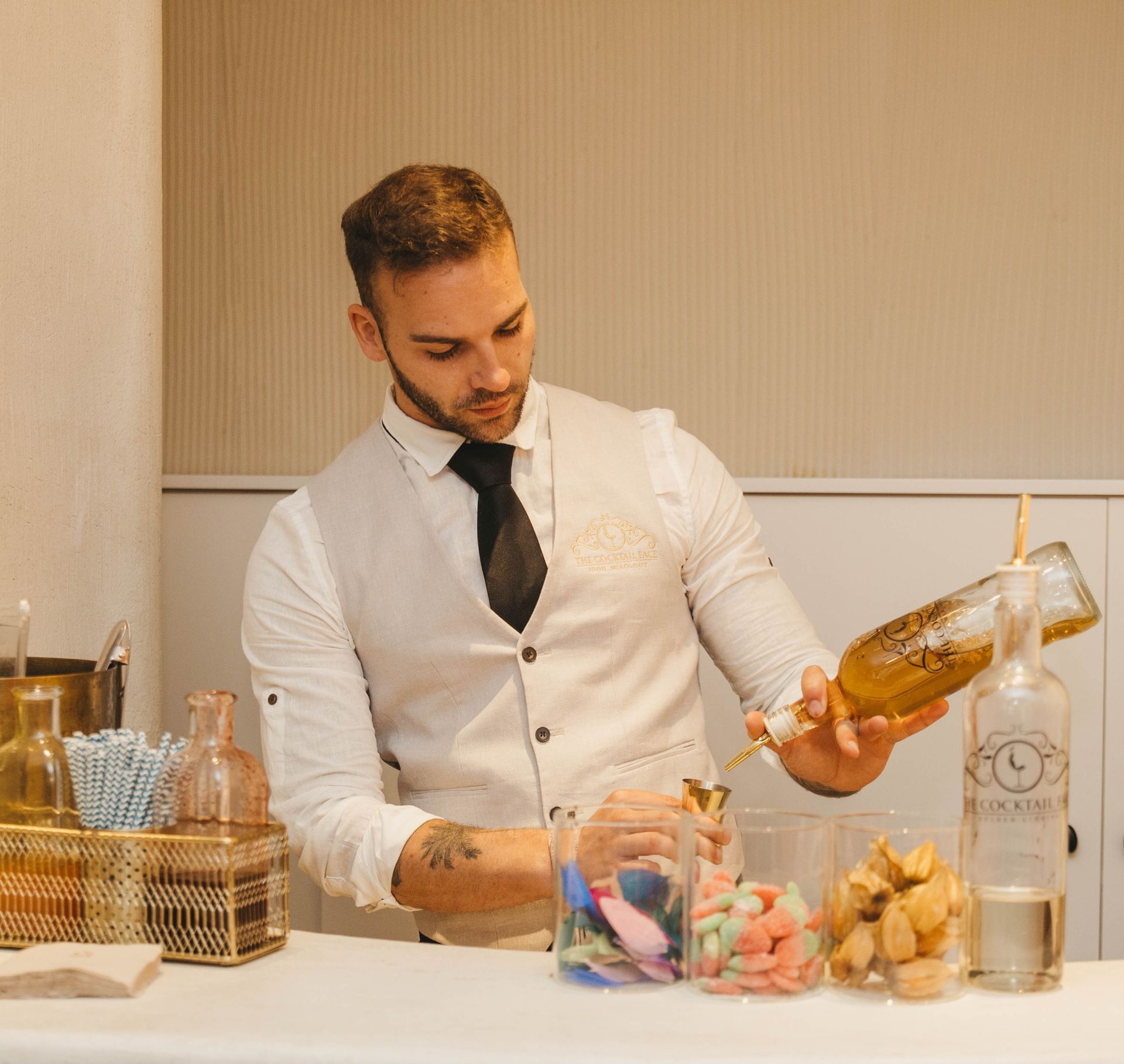 Camarero con chaleco blanco sirviendo de una botella detrás de la barra; cristalería y aperitivos delante.