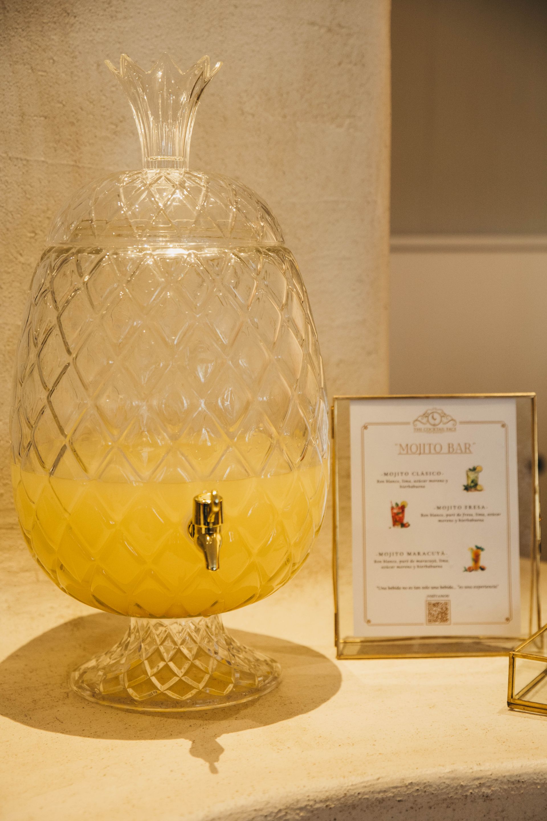 Dispensador de bebidas de cristal con forma de piña y líquido amarillo. Un cartel al lado dice