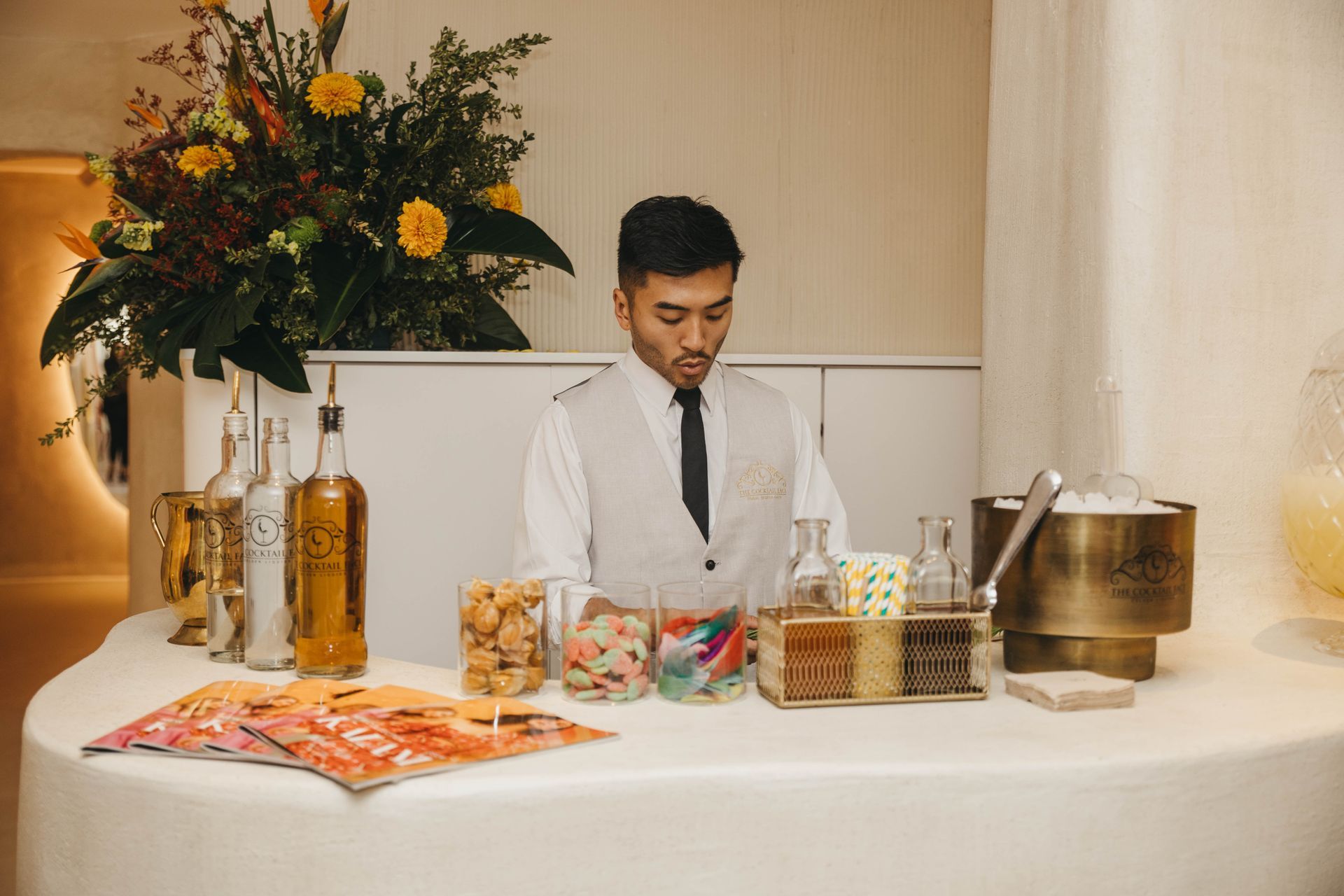 Un camarero prepara bebidas sentado en una barra blanca curva. Hay flores detrás de él.