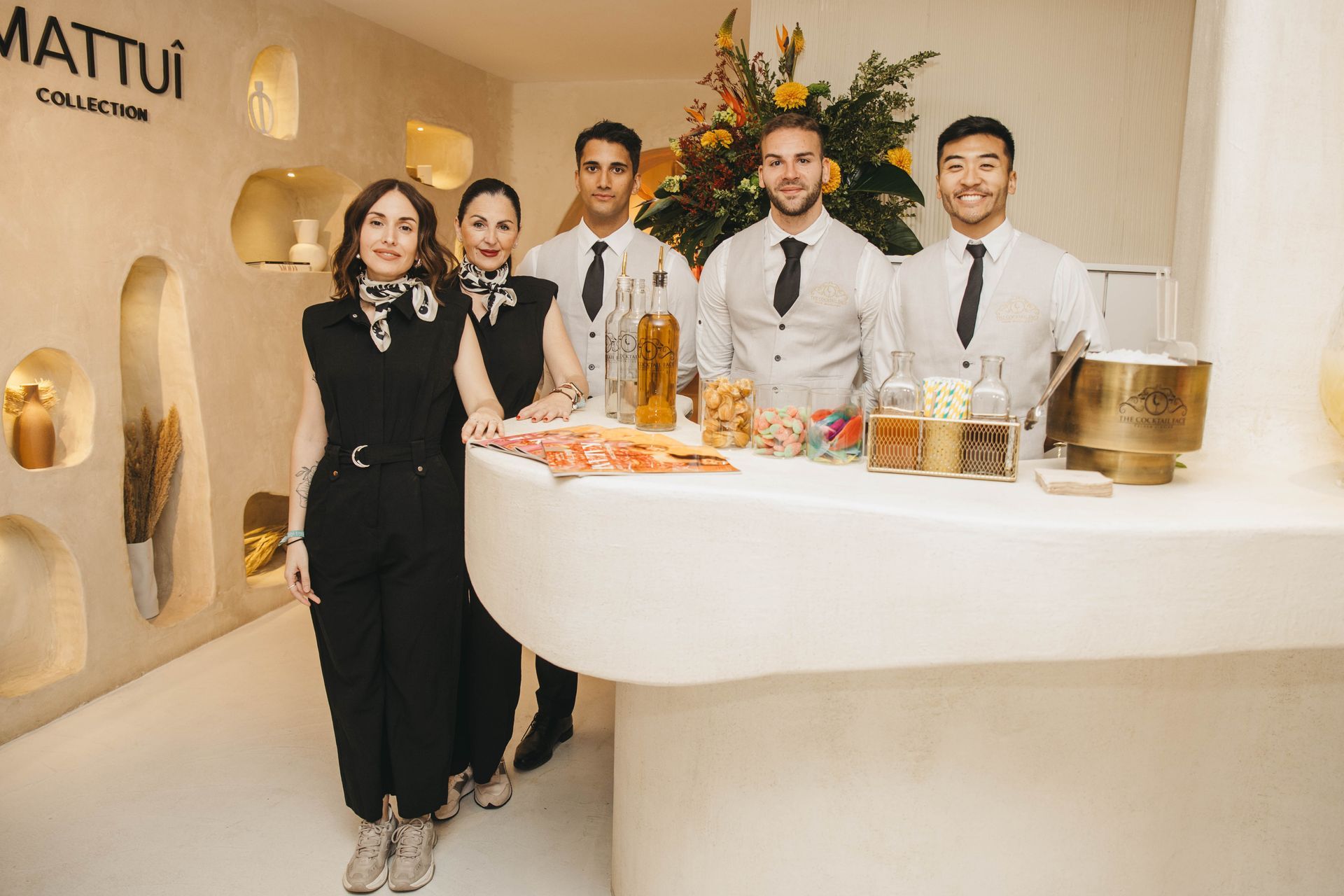 Seis personas están de pie detrás de una barra blanca con aperitivos y bebidas, dentro de una habitación de tonos claros.
