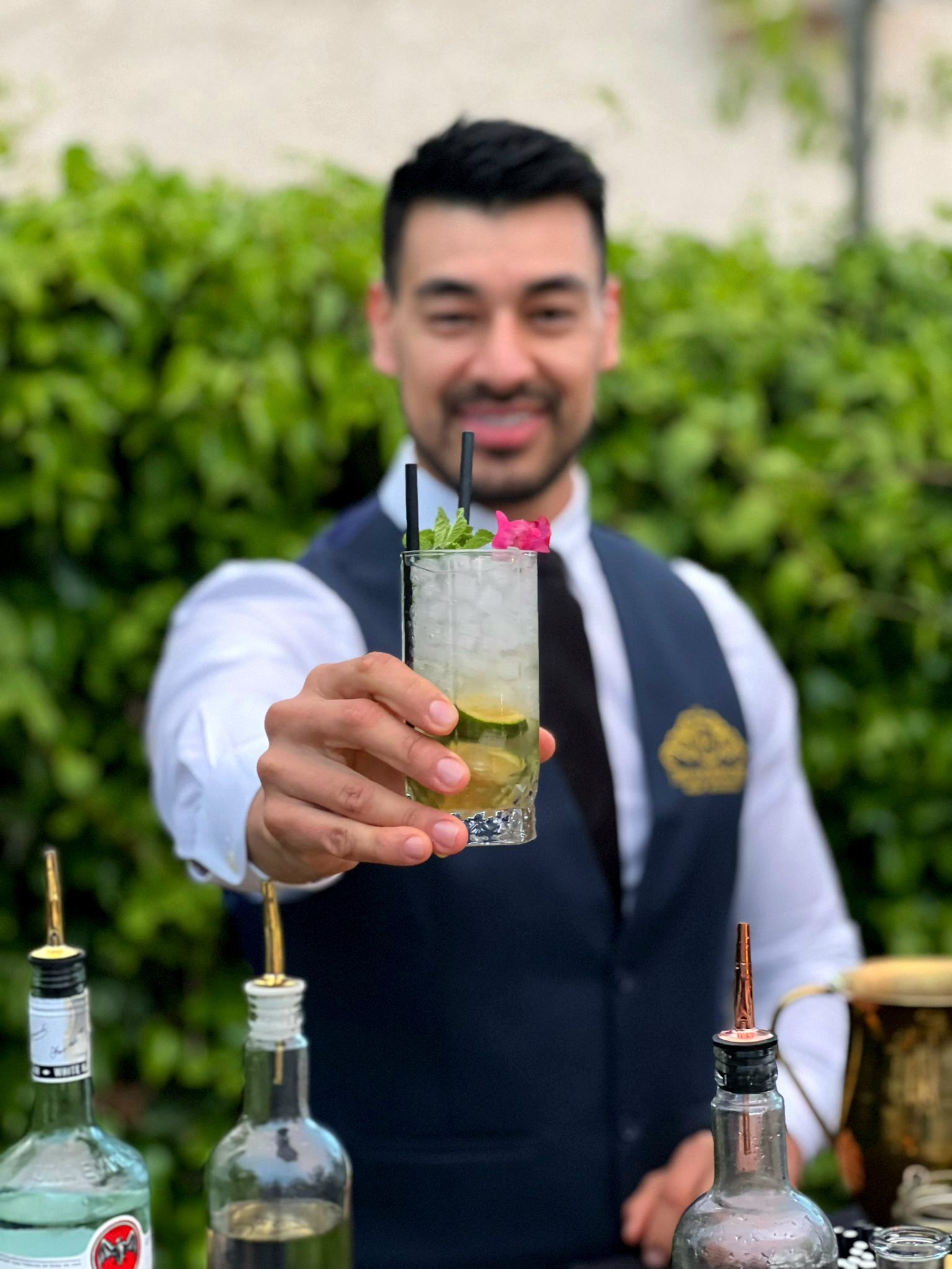 Un camarero sonriente, con un cóctel en la mano, está delante de una barra exterior con botellas.