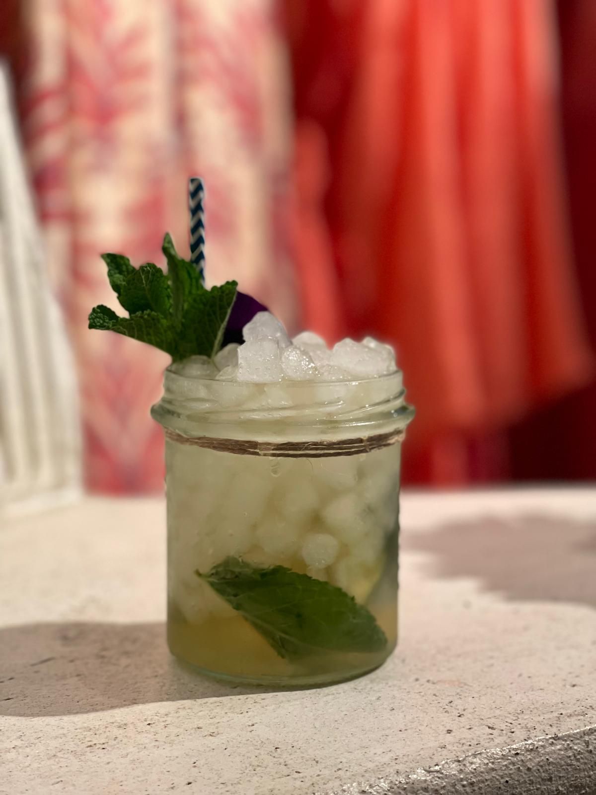 Cóctel en tarro de cristal con hielo picado, adorno de menta, pajita, sobre un fondo de ropa desenfocada.