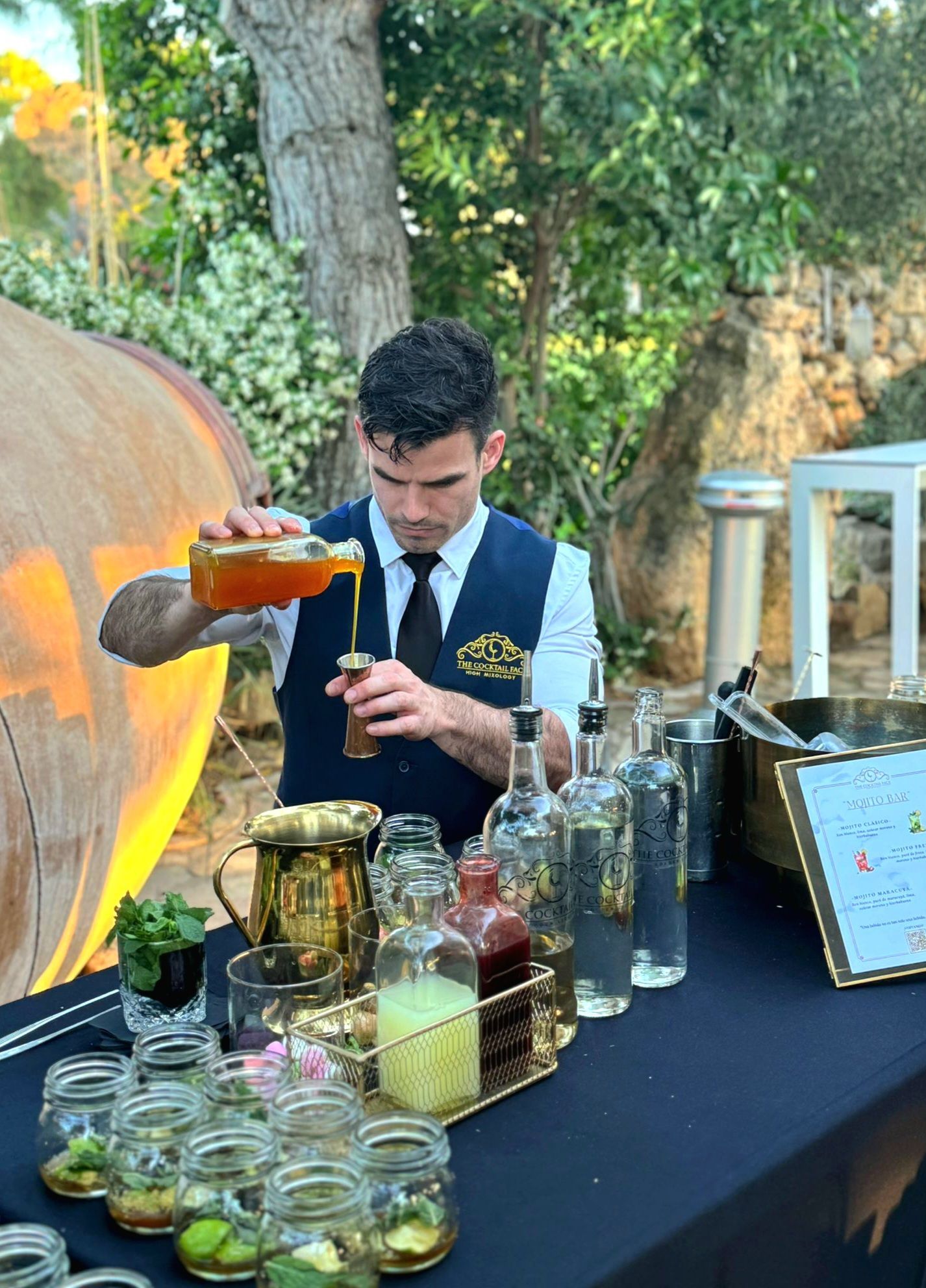 Un camarero sirve una bebida en un bar al aire libre con botellas, tarros y un barril.