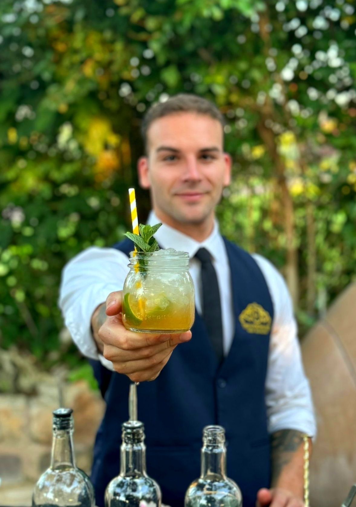 Camarero con cóctel en mano, sonriendo; ambiente al aire libre, camisa blanca, chaleco azul, detalles dorados.