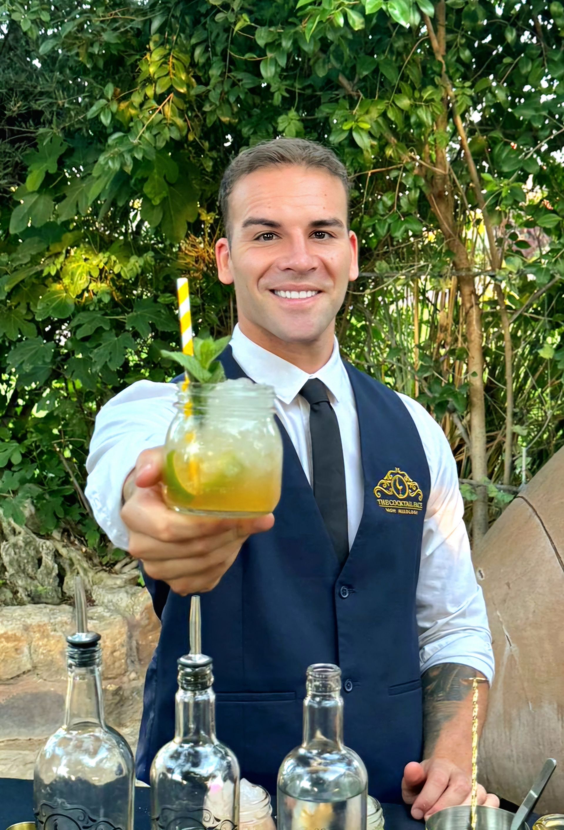 Camarero con un cóctel, sonriendo. Vestía chaleco azul y camisa blanca.