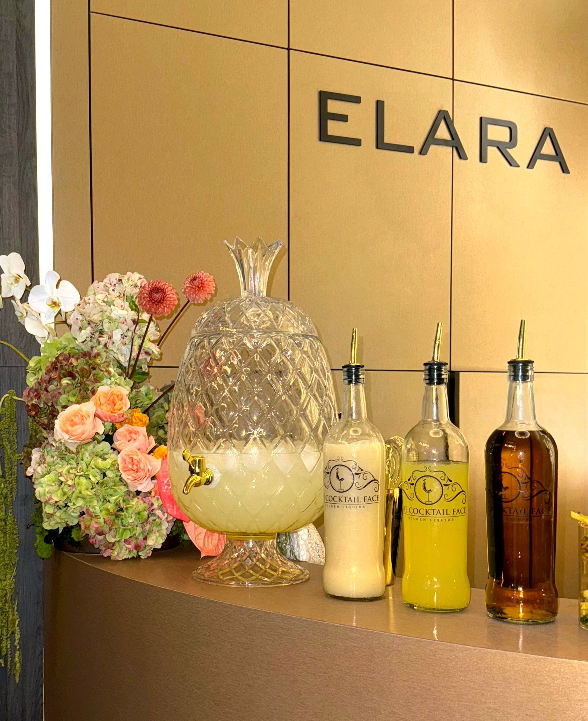 Bar montado con dispensador de limonada, botellas de bebidas y flores delante de un cartel que dice