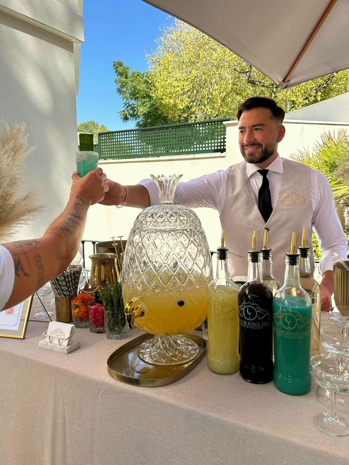 Un camarero en un bar al aire libre, sonriendo y brindando con un cliente, cerca de un gran dispensador de bebidas.
