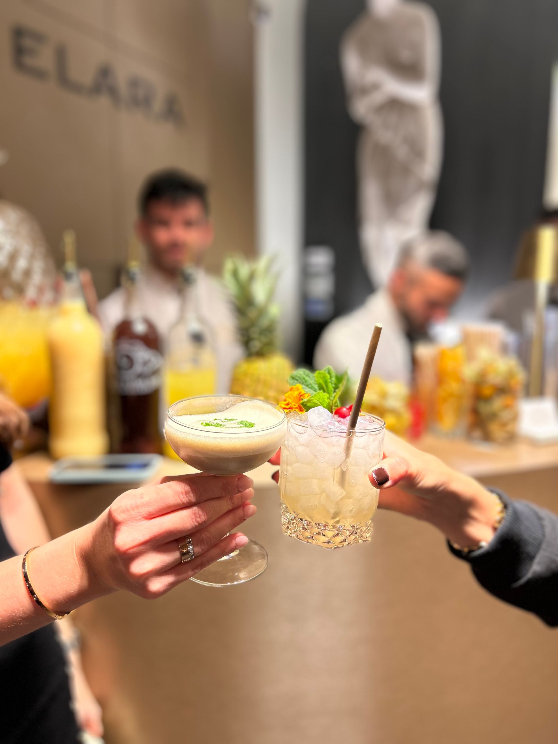 Dos manos chocan cócteles en un bar. Al fondo, camareros; una piña y otras bebidas visibles.