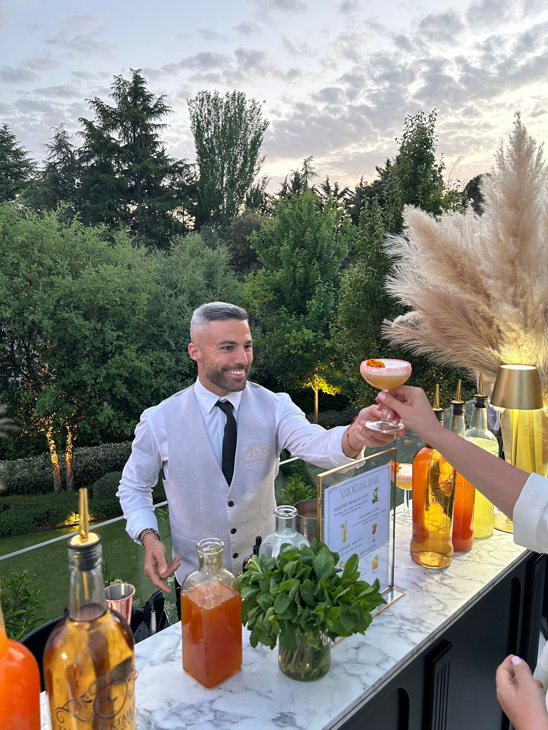 Camarero sirviendo cóctel en un bar al aire libre con árboles al fondo.