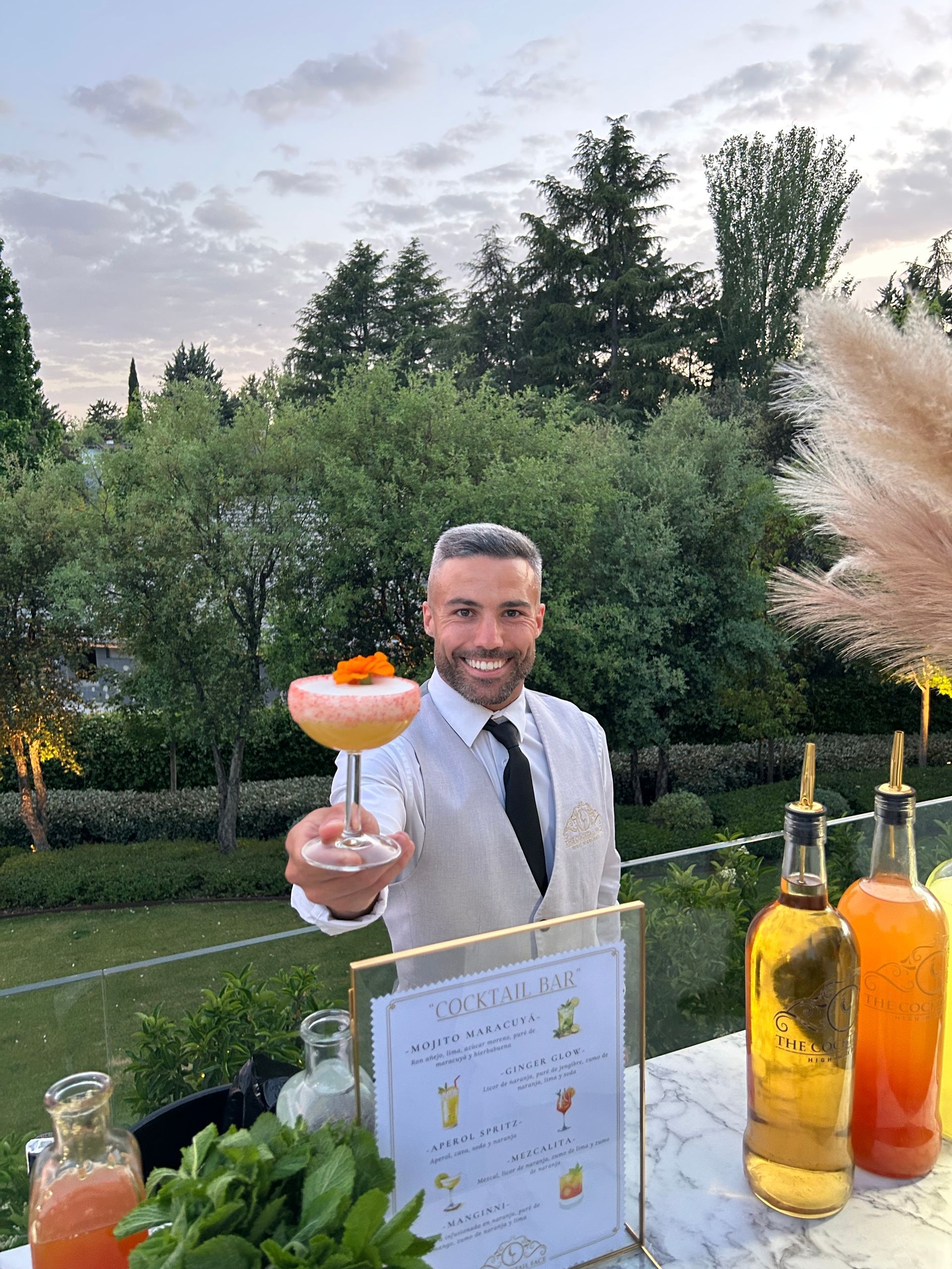 Camarero sonriente con un cóctel al aire libre. Menú visible.