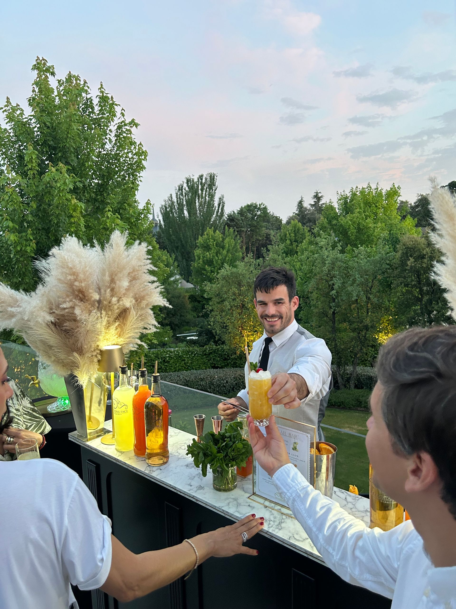Un camarero sirve un cóctel a una persona en un bar al aire libre, sonriendo. Atardecer con vegetación al fondo.
