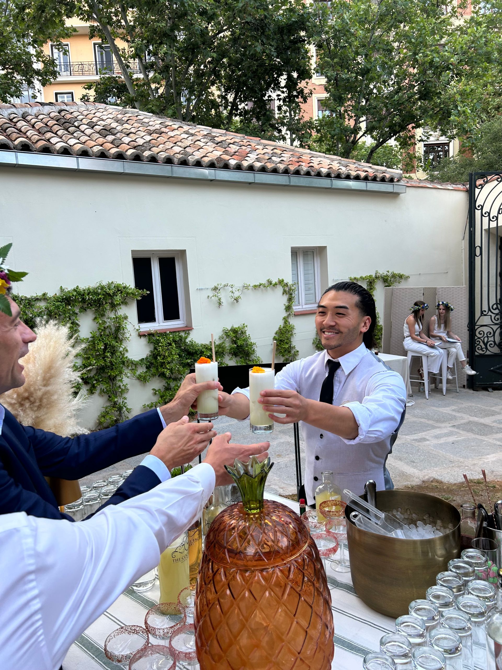 Camarero sirviendo cócteles; invitados brindando. Exterior: piña dorada, cubo de hielo y edificio blanco.