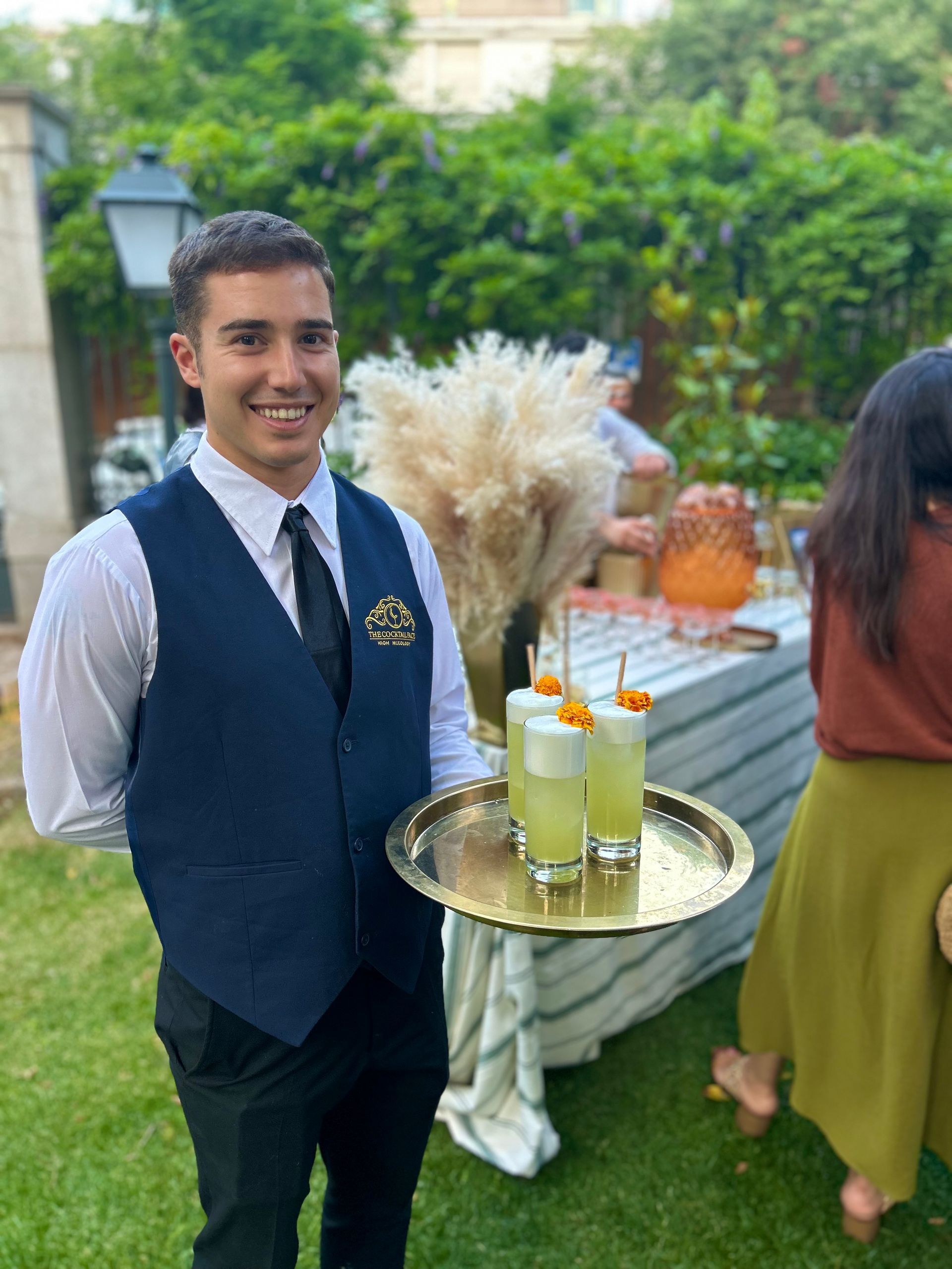 Camarero sonriendo con una bandeja de tres cócteles. Evento al aire libre, con una estación de bebidas al fondo.