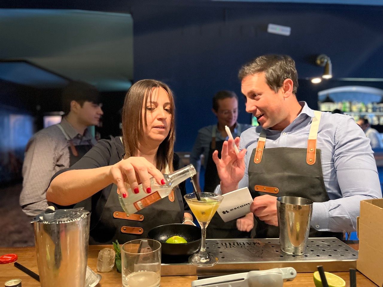 Dos personas preparan cócteles tras una barra. Una sirve de una botella, la otra gesticula. Al fondo, se ven otras personas y la barra.