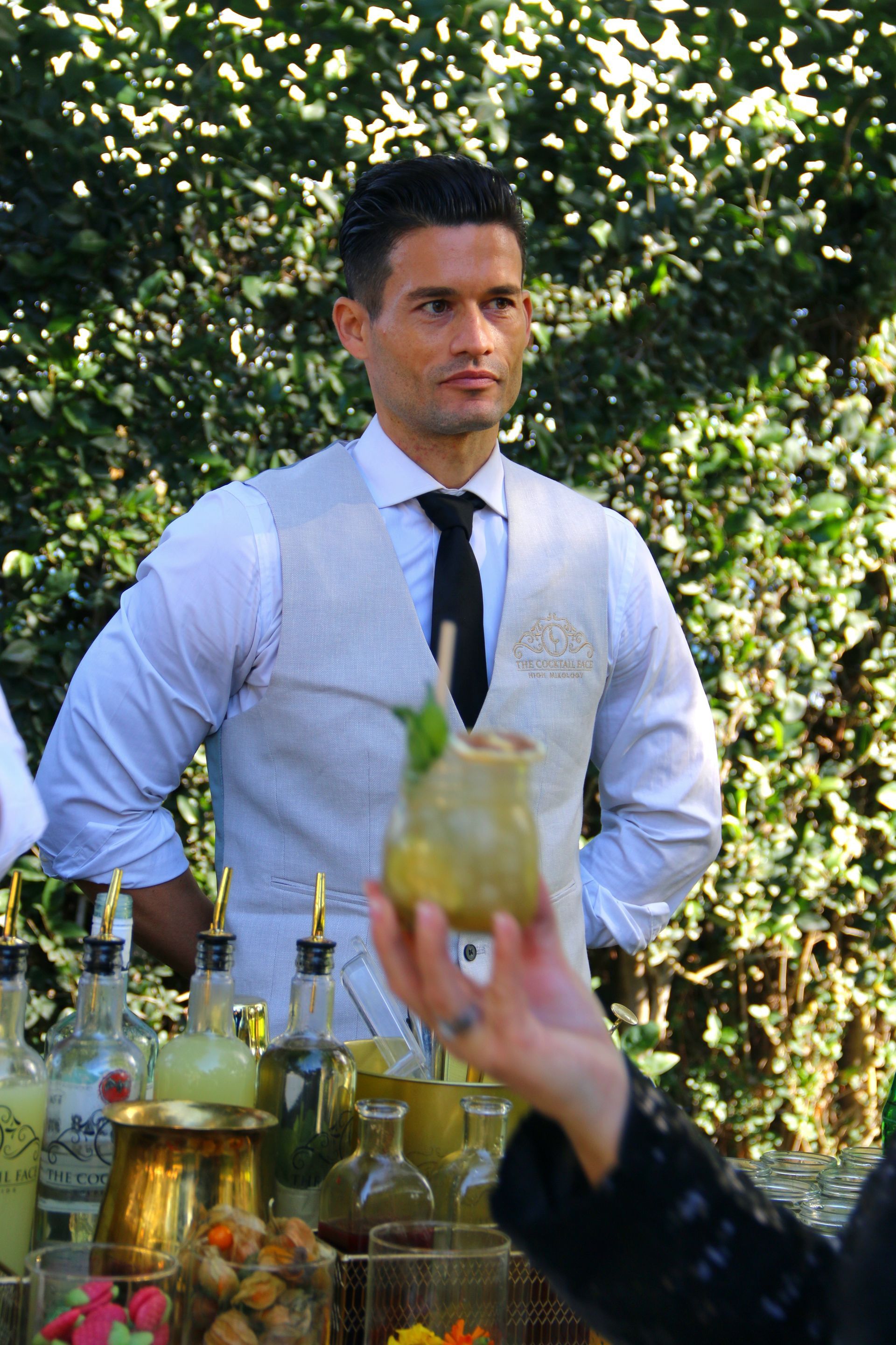 Hombre con chaleco y corbata, detrás de una barra, con cóctel en mano. En un entorno al aire libre con vegetación.