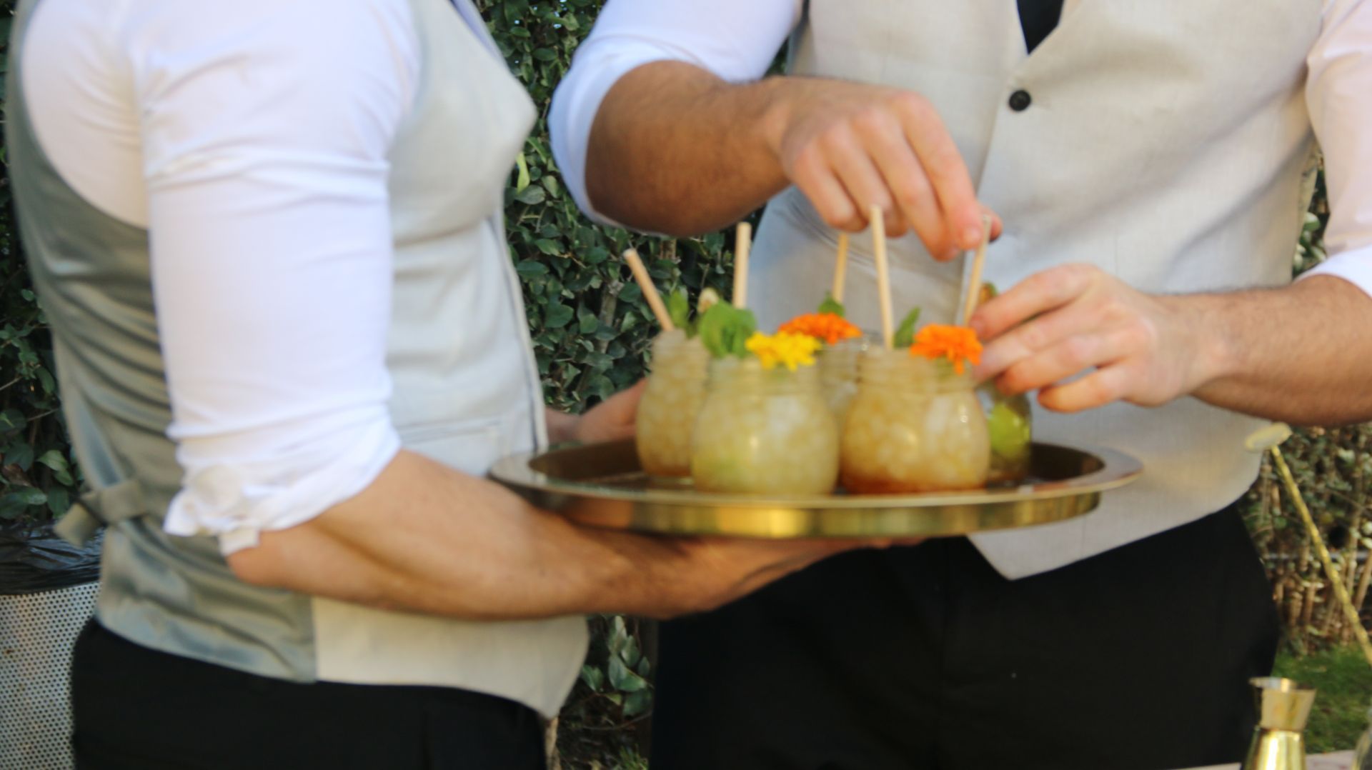 Dos personas sirviendo aperitivos en una bandeja dorada al aire libre, añadiendo guarniciones.