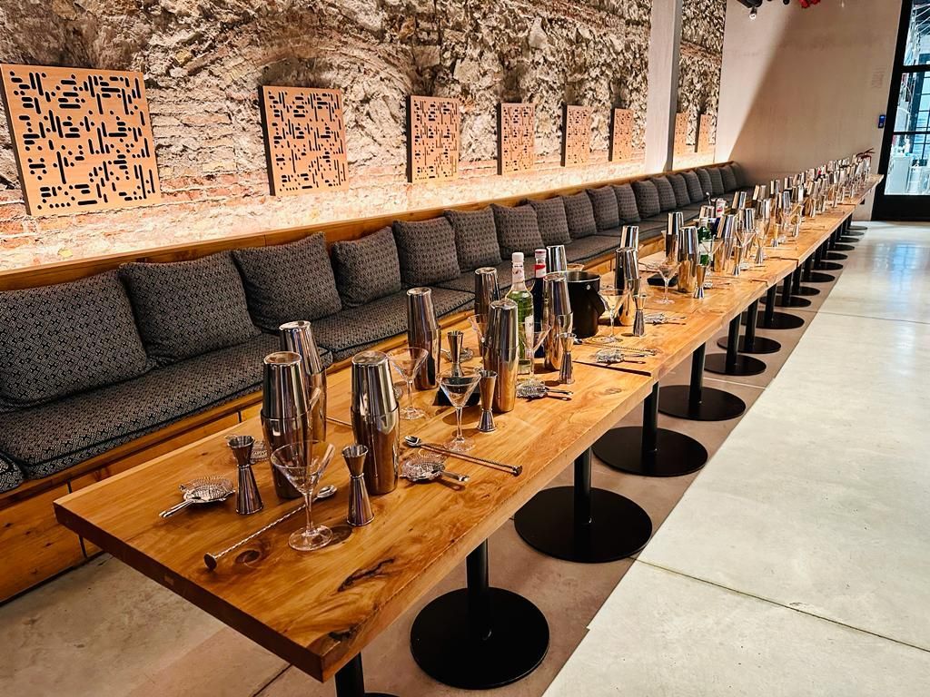 Una larga mesa de madera con cocteleras y cristalería, junto a un banco acolchado.