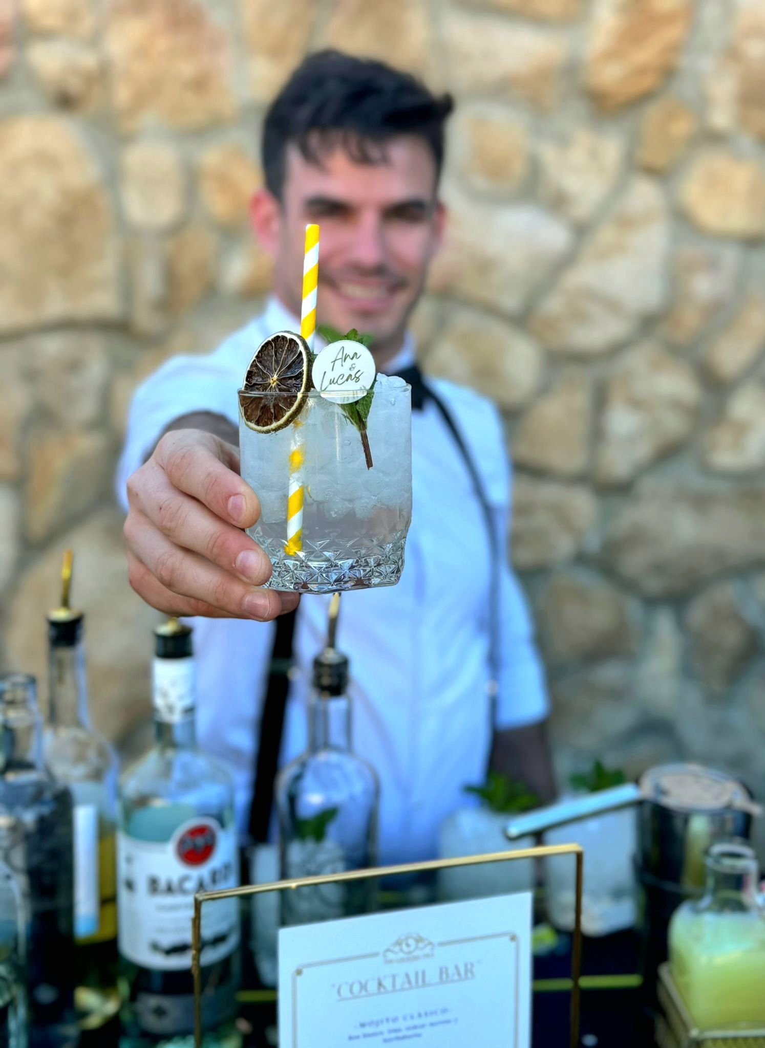 Un camarero sonríe mientras sostiene un cóctel con pajita y adornos. En la barra hay botellas y un cartel.