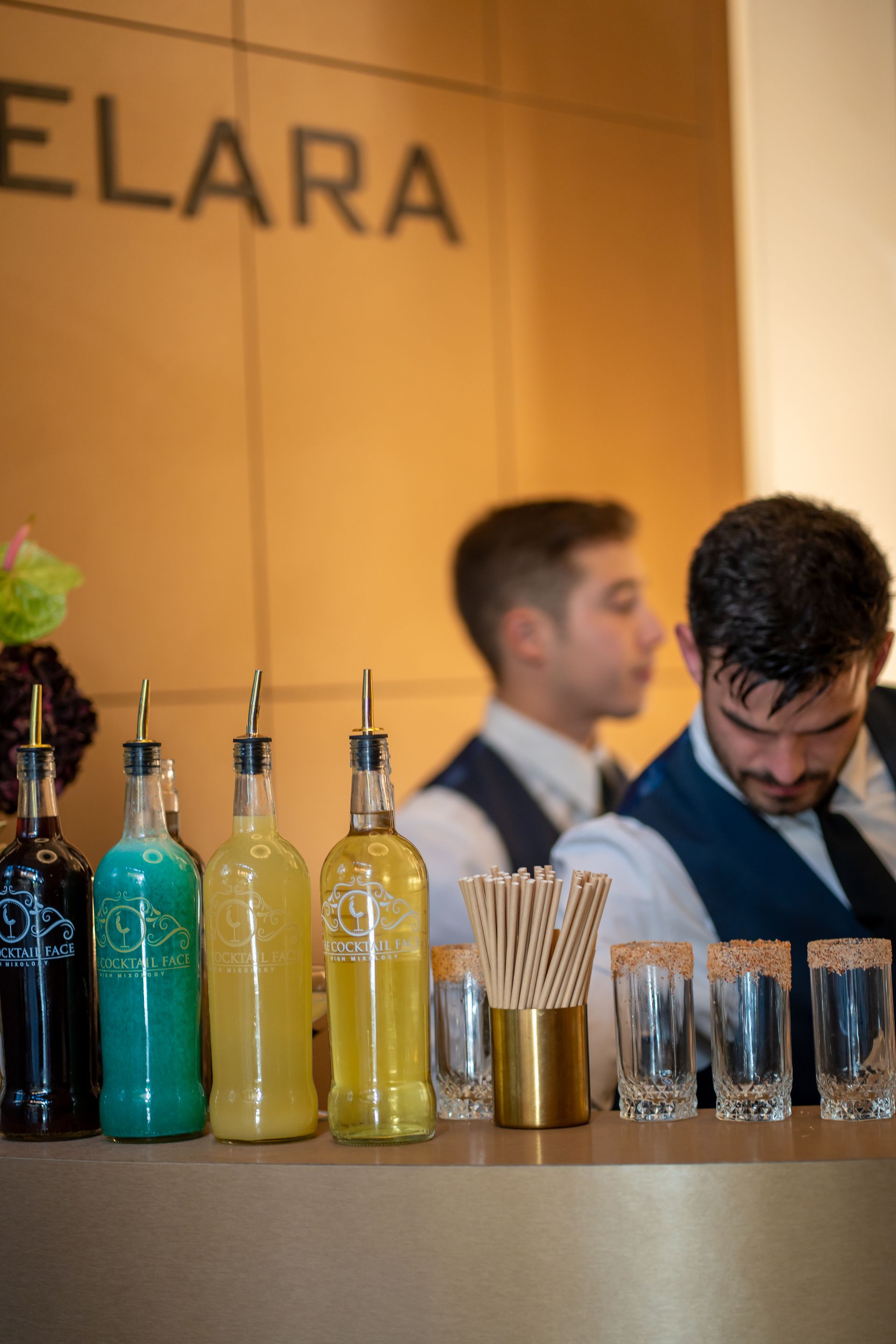 Bar con botellas de siropes de colores, camareros y copas de cóctel.