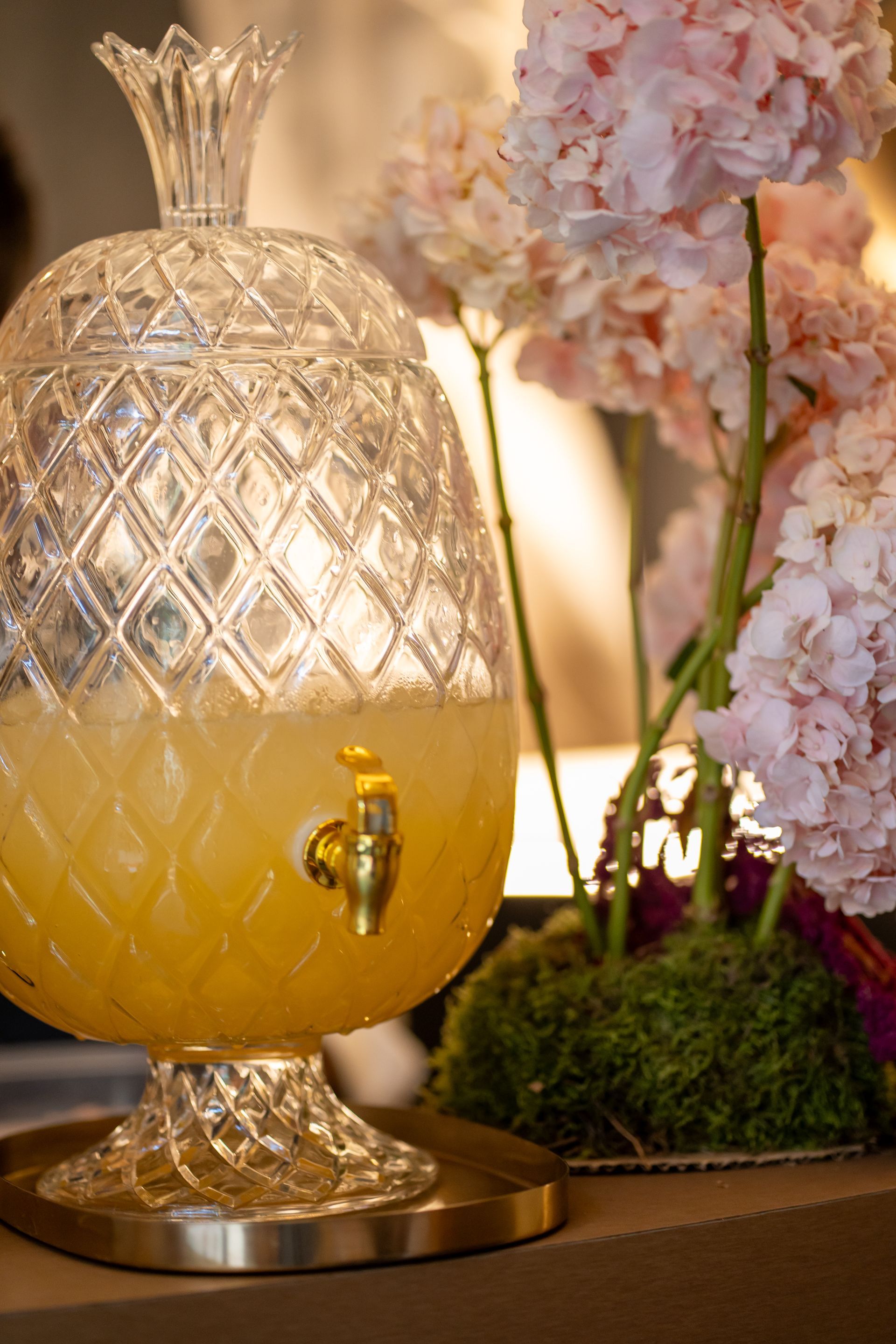 Dispensador de bebidas de cristal con forma de piña, lleno de líquido amarillo, junto a flores rosas sobre musgo.