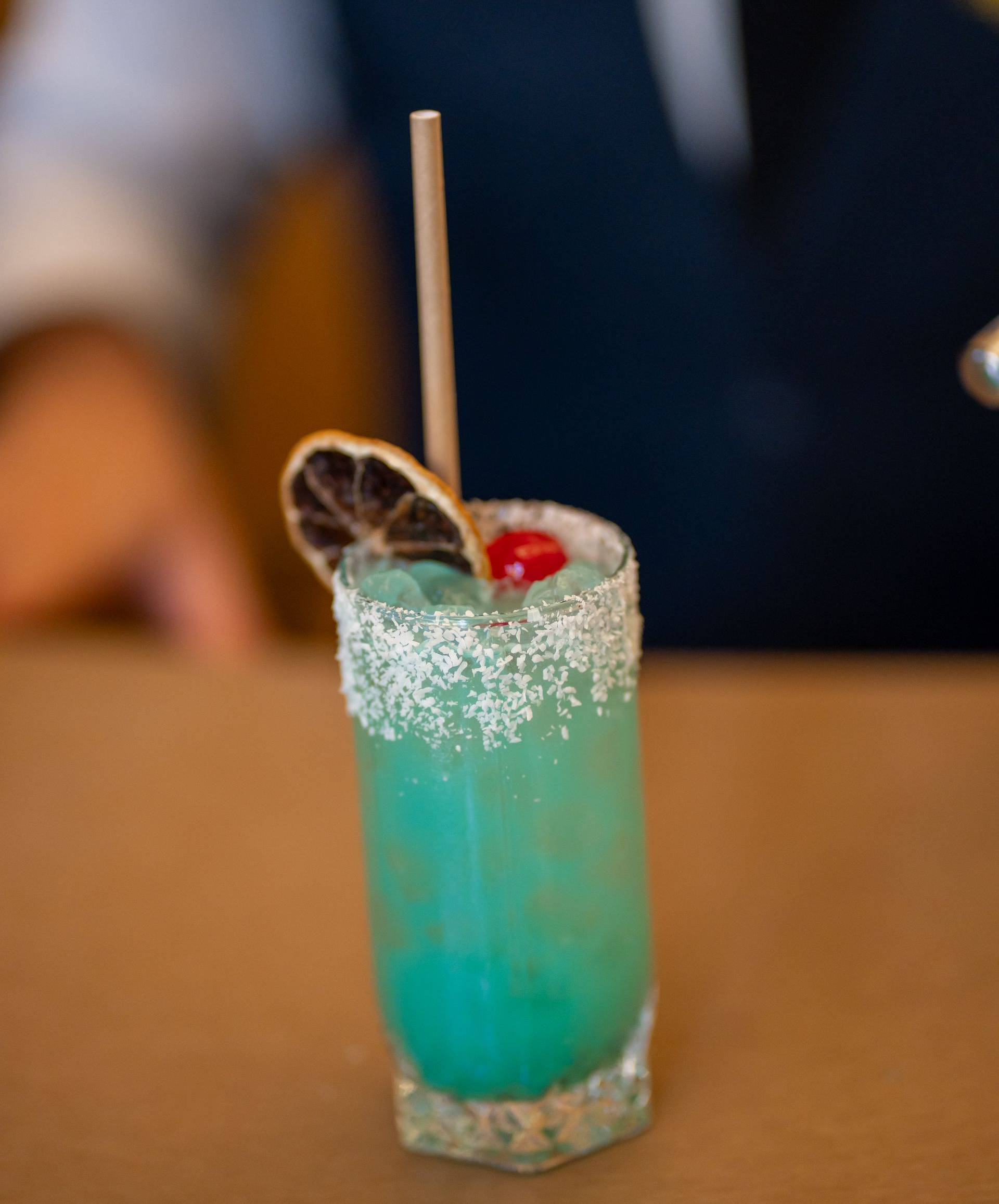 Cóctel azul servido en un vaso con borde de azúcar, adornado con una cereza y una rodaja de naranja deshidratada, con una pajita.