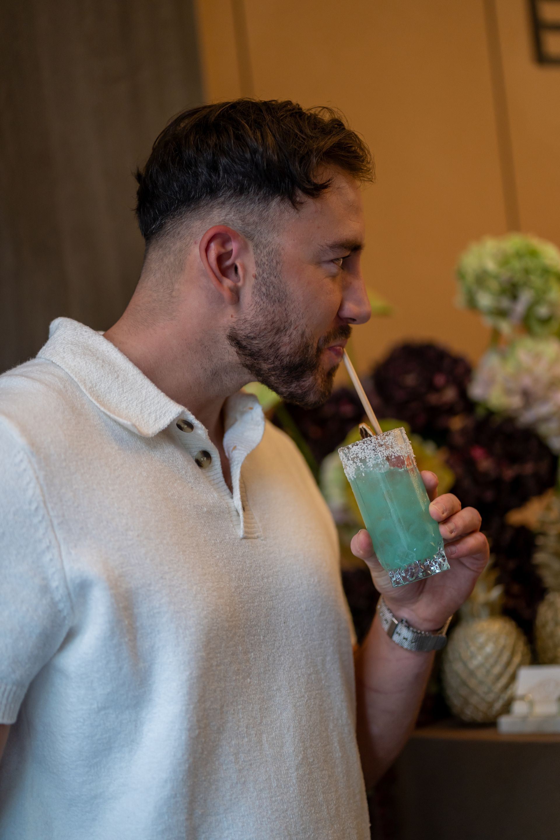 Hombre bebiendo un cóctel azul con pajita. Viste un polo blanco y se encuentra en un espacio bien iluminado.