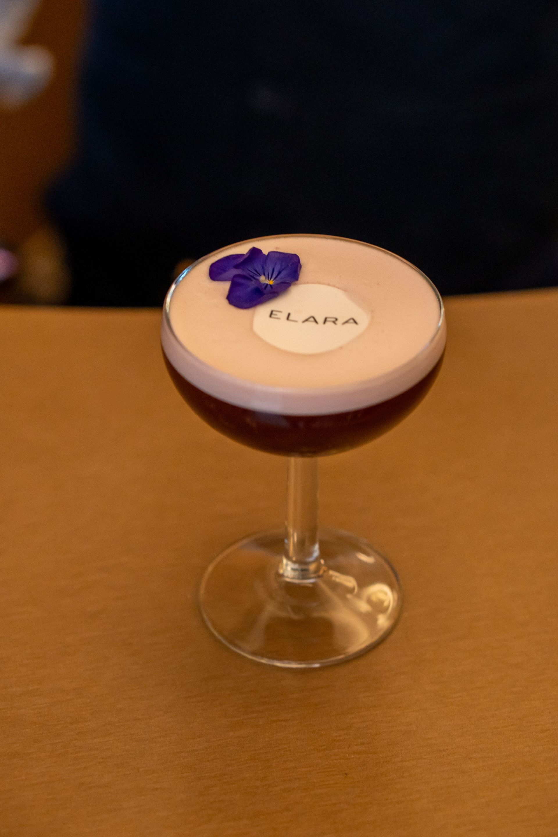 Cóctel servido en copa de pie, con espuma de lavanda, base de color púrpura oscuro, adorno floral y la palabra