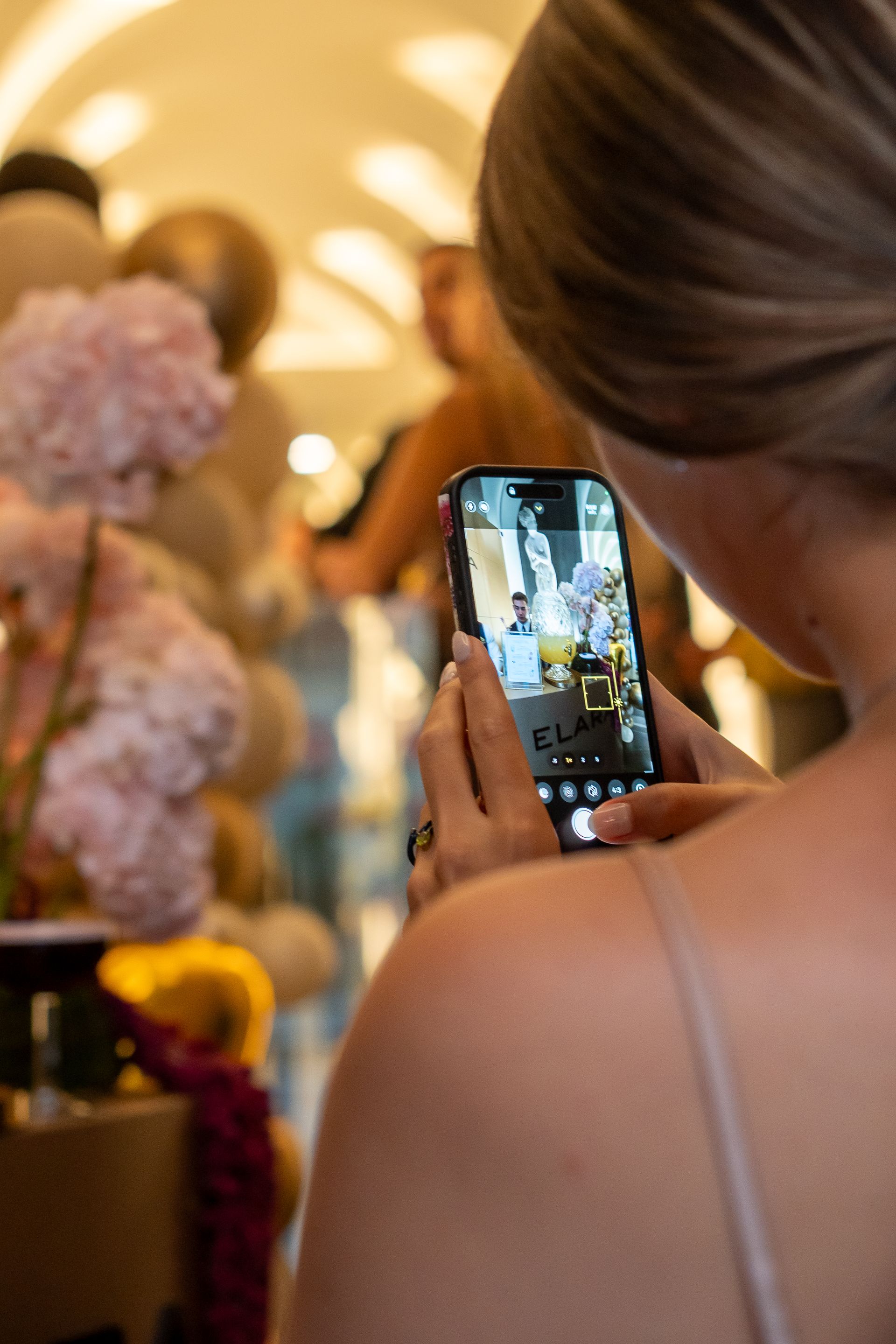 Mujer fotografiando un evento con su teléfono inteligente. Flores rosas, globos y otras personas aparecen al fondo desenfocado.