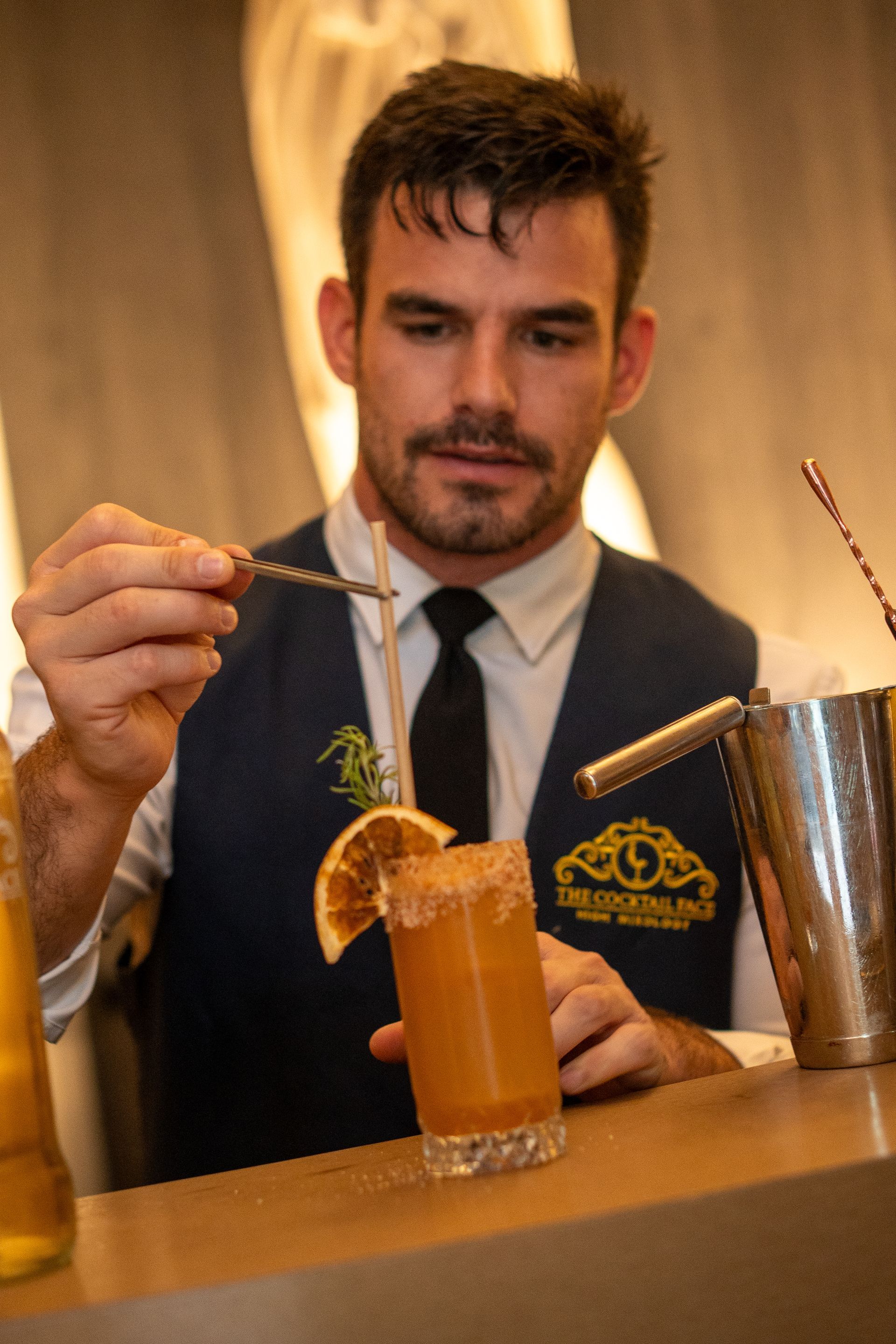 El camarero añade un adorno al cóctel: una rodaja de naranja y una ramita de romero en el borde de la copa. Lleva chaleco y corbata; el ambiente es el de un bar.