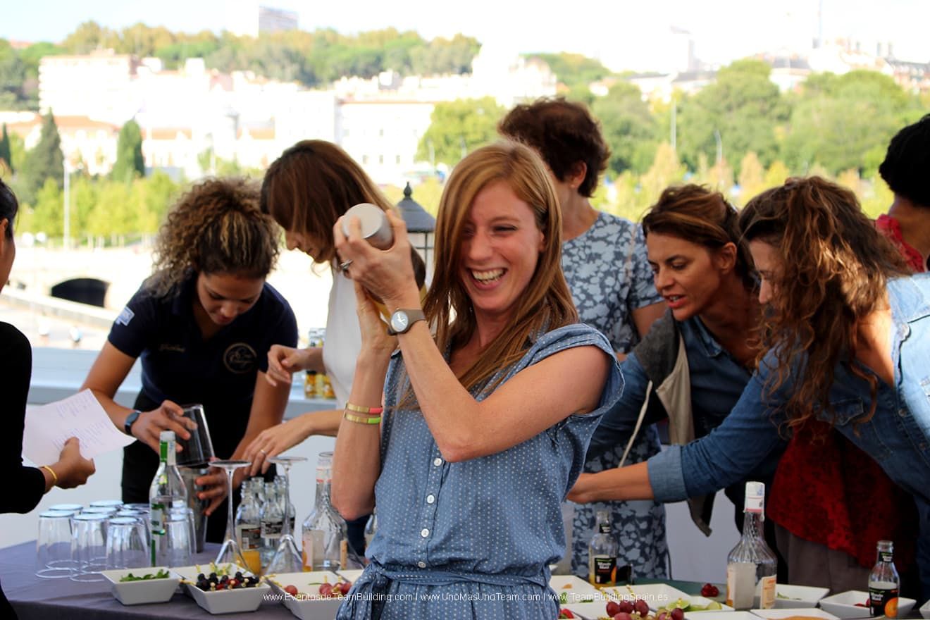 Mujer agitando una coctelera, riendo, en un evento al aire libre, rodeada de otras personas, con fruta y botellas sobre la mesa.