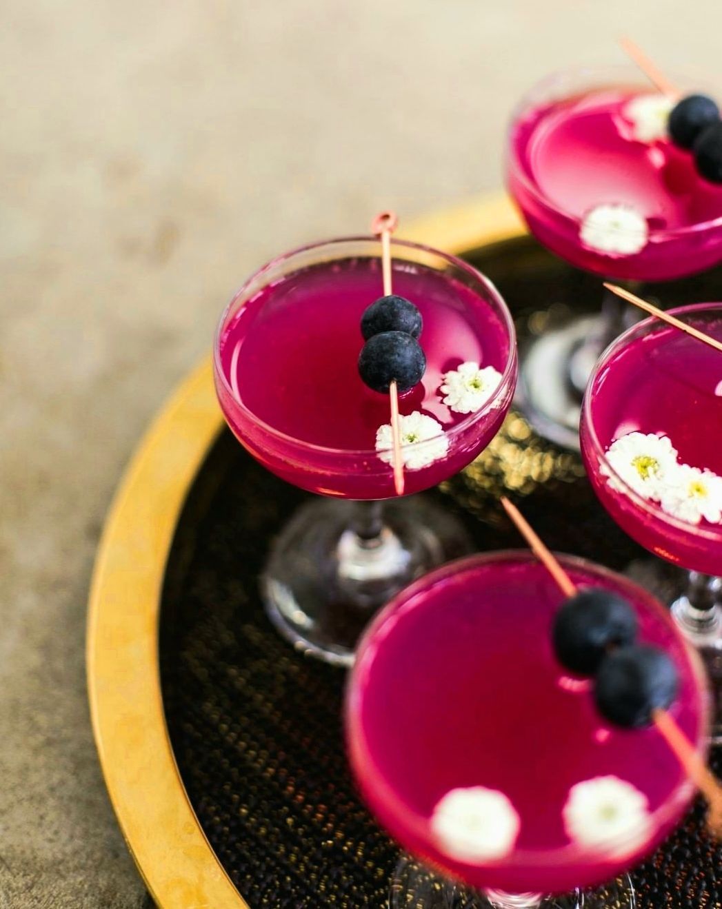 Cuatro cócteles magenta adornados con arándanos y flores blancas en una bandeja.