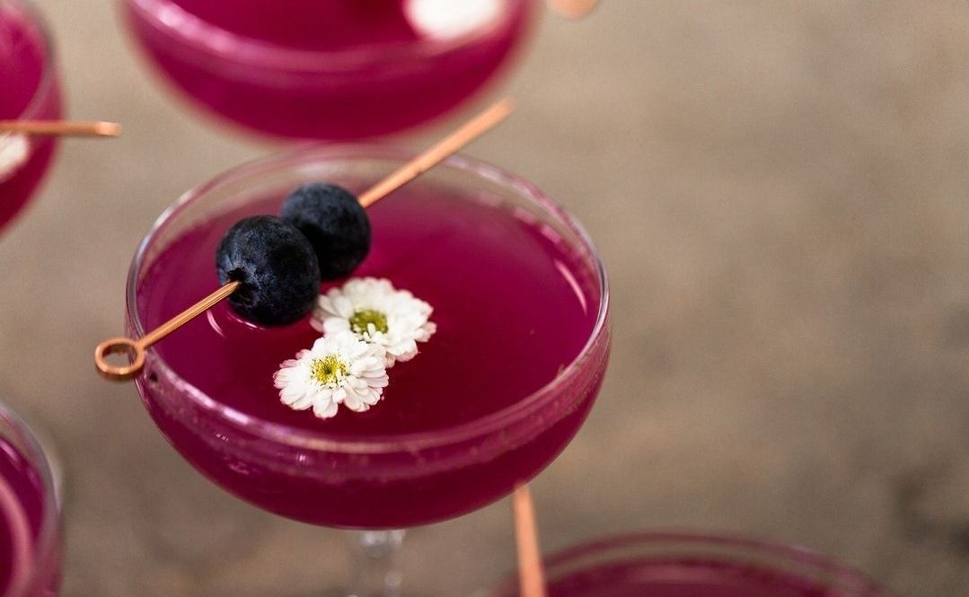 Cócteles morados adornados con arándanos y flores blancas, sobre una mesa.