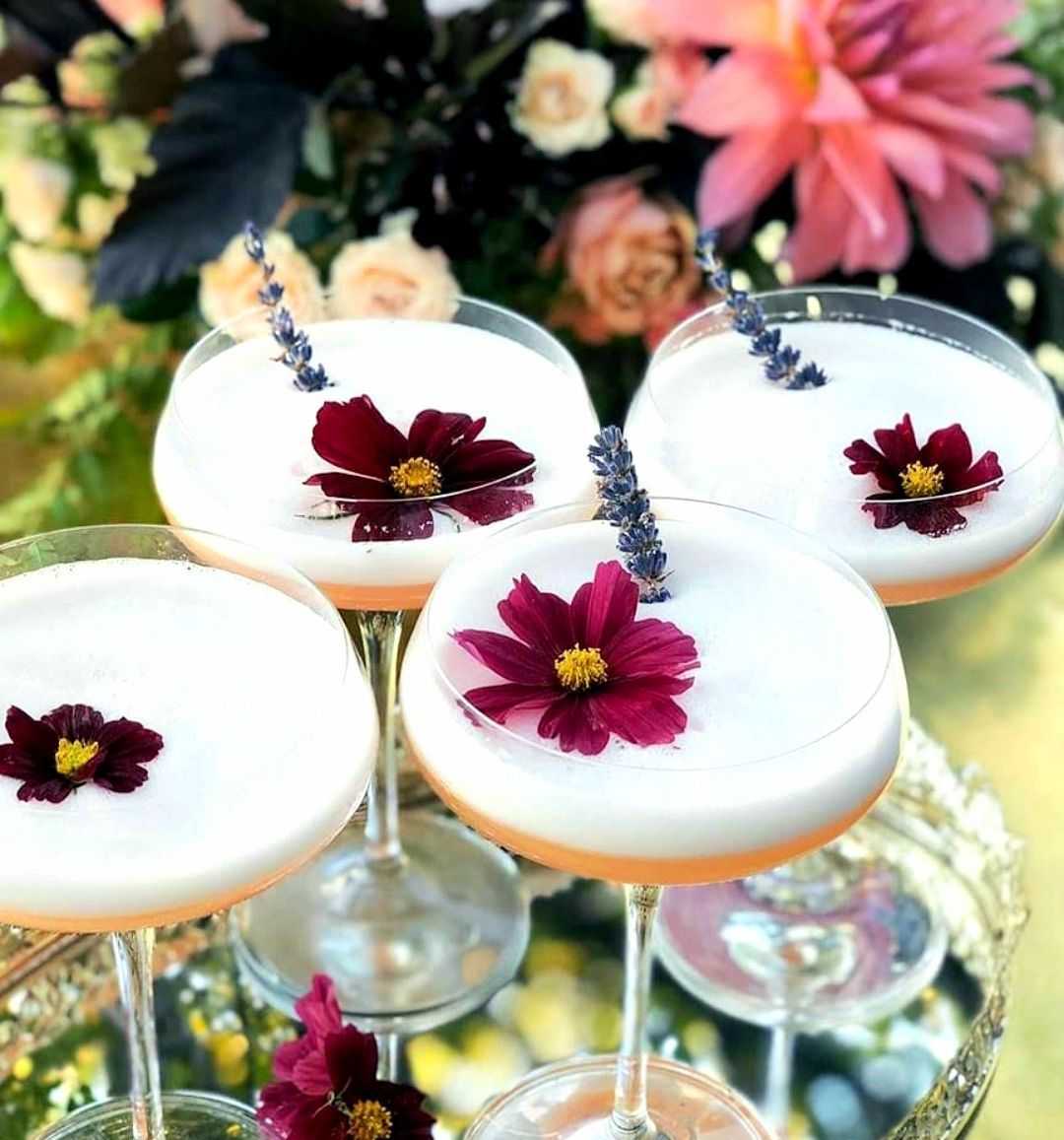 Cuatro cócteles rosados, adornados con flores, reposan en una bandeja de plata frente a un arreglo floral.