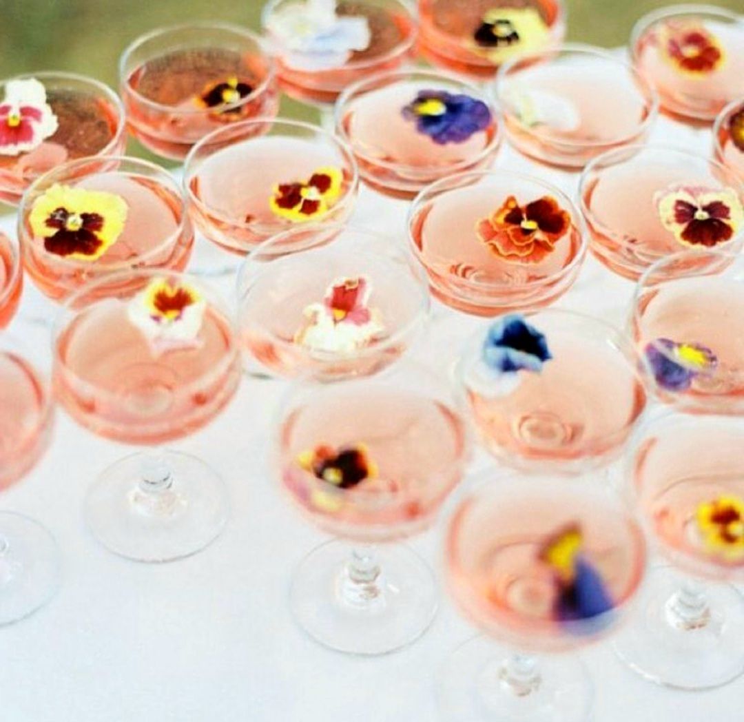 Copas de cócteles rosados, cada una adornada con flores comestibles, dispuestas sobre una superficie blanca.