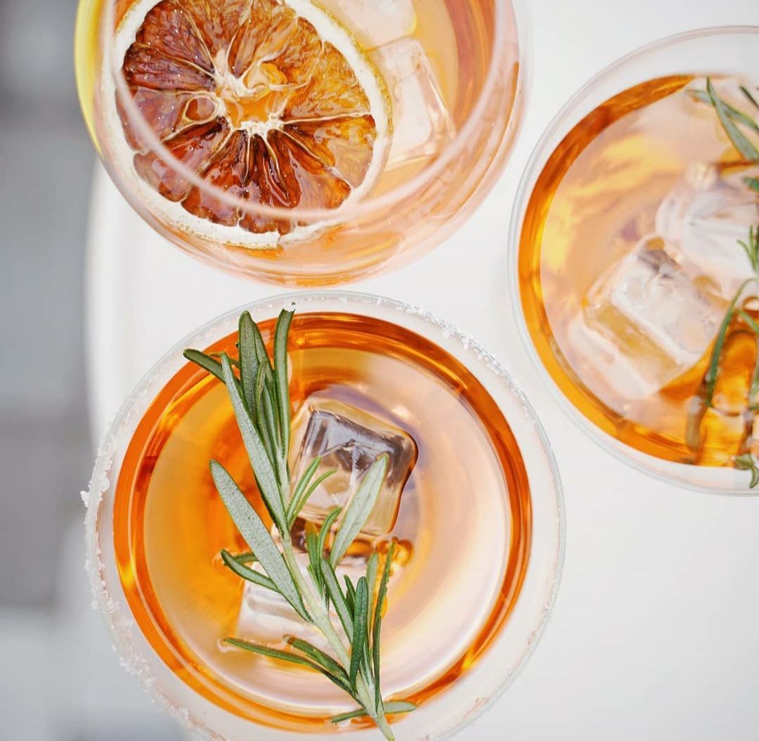 Tres cócteles, de color ámbar con hielo, romero y rodajas de naranja, sobre una superficie blanca.