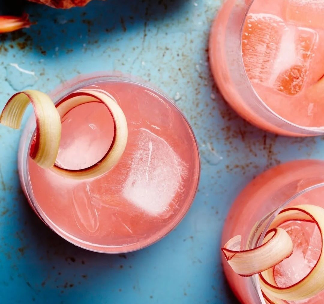 Tres cócteles rosas con cubitos de hielo y guarnición de cáscara de manzana sobre una superficie azul.