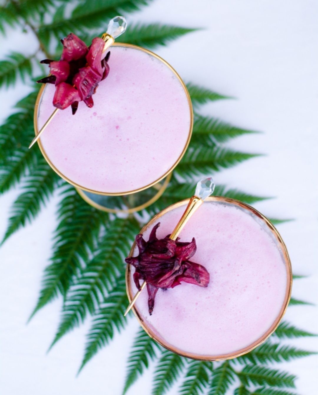 Dos cócteles rosas con guarnición de hibisco sobre un helecho.