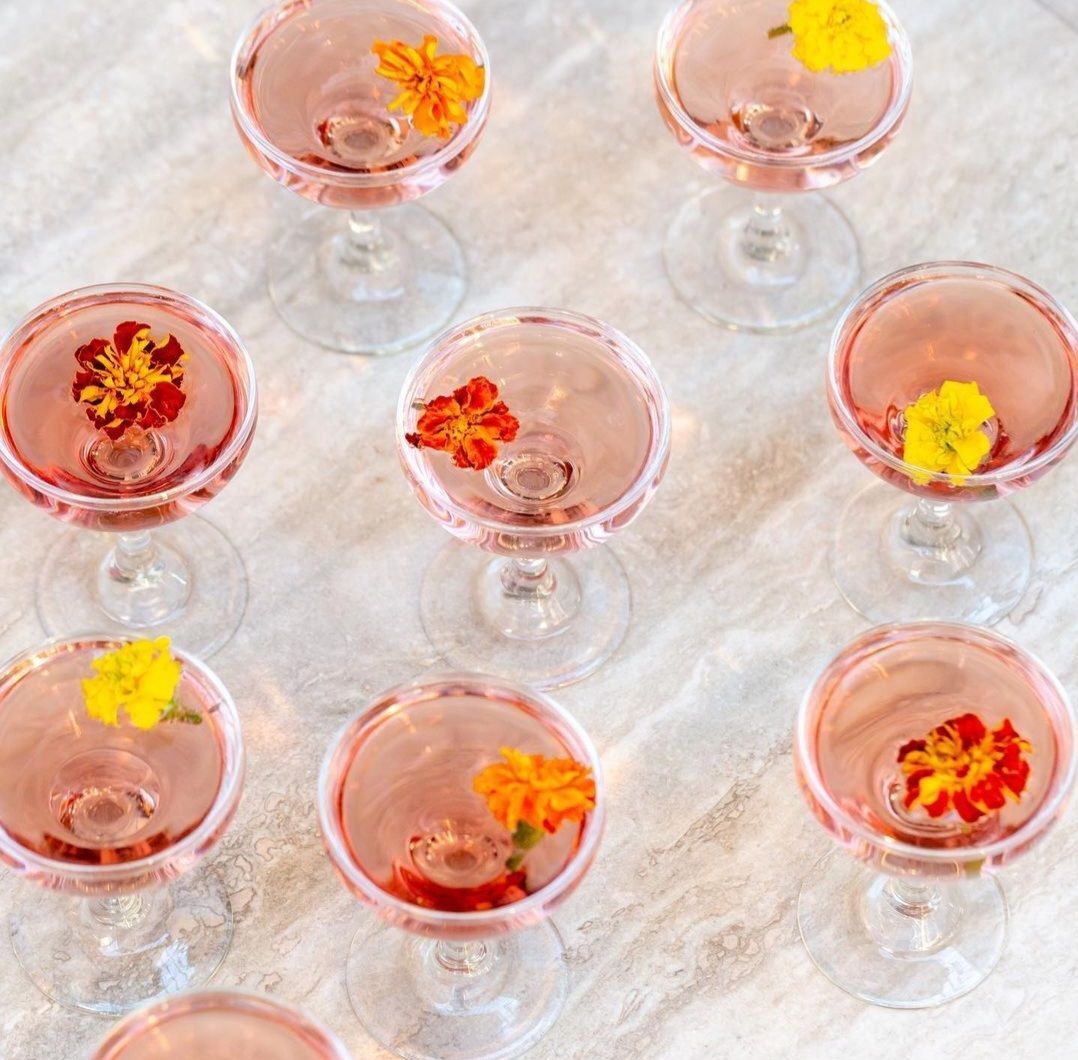 Nueve cócteles rosas en copas coupé, cada uno con una guarnición de flores sobre una superficie de mármol claro.