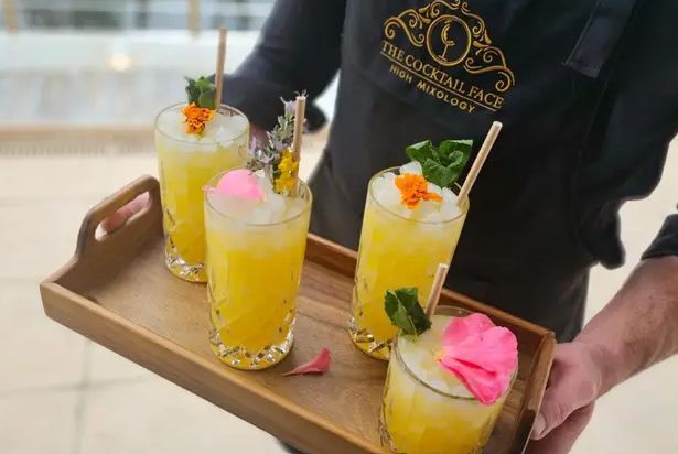 Camarero sosteniendo una bandeja de cuatro cócteles amarillos con hielo, guarniciones y pajitas.