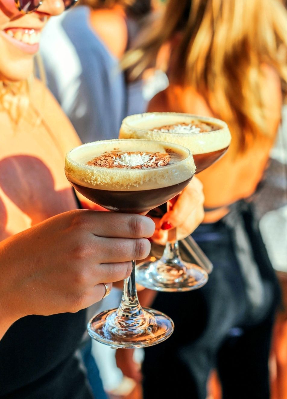 Dos martinis espresso sostenidos por una persona con cara sonriente, al aire libre, con fondo borroso.