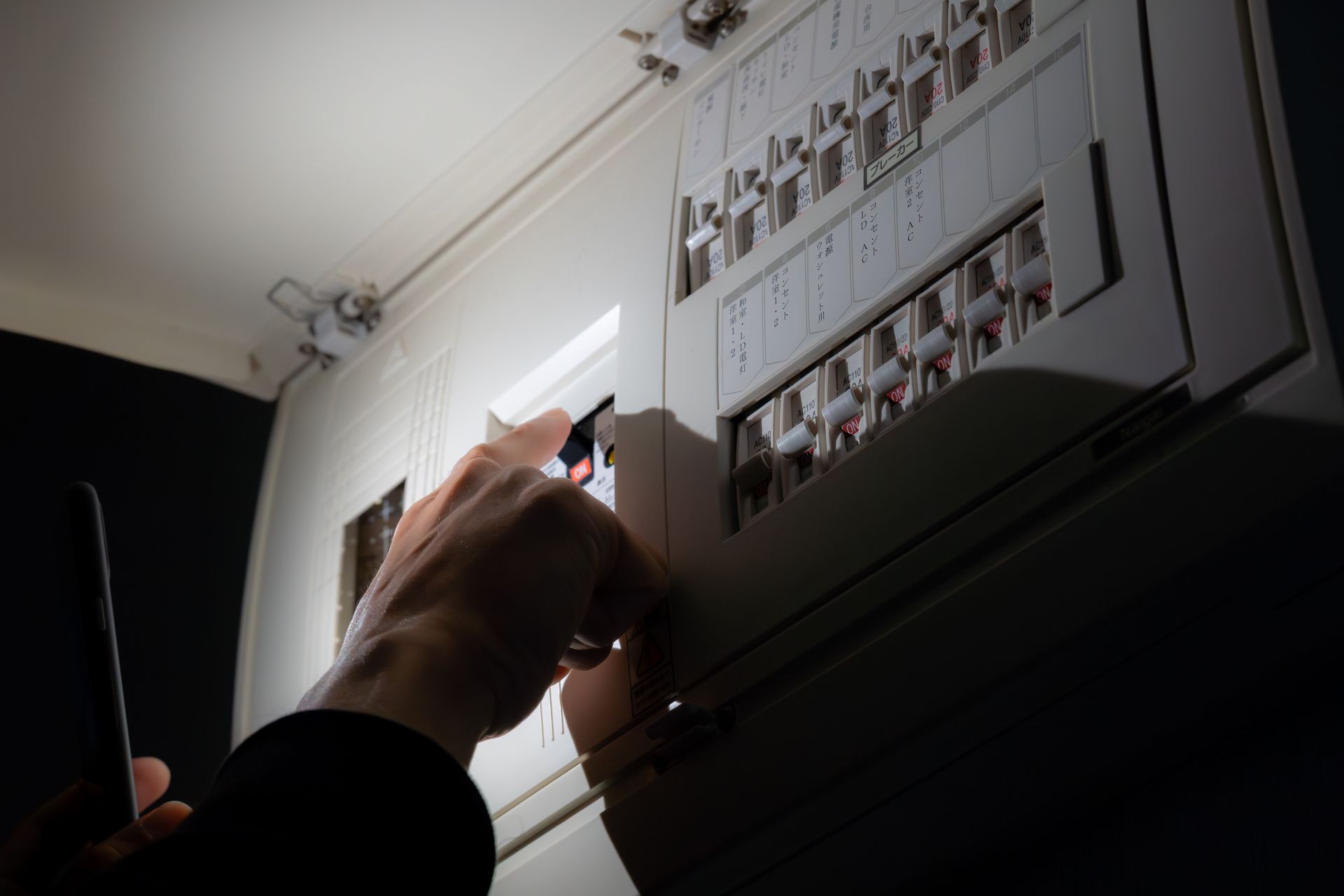 Une personne utilise la lampe de son téléphone pour inspecter un tableau électrique résidentiel dans une pièce faiblement éclairée.