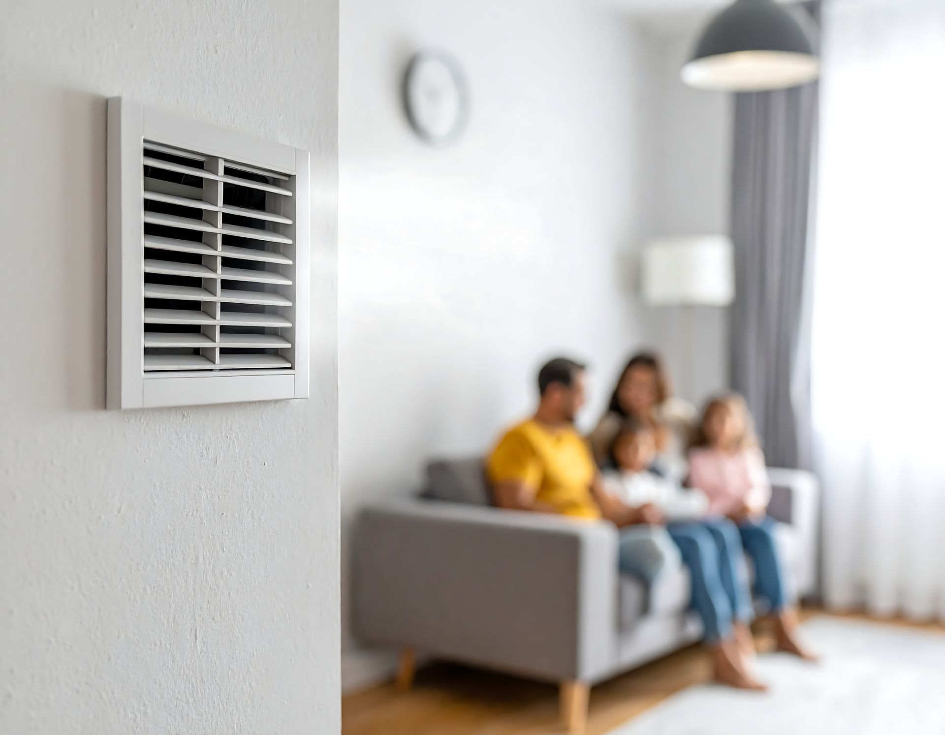 Au premier plan, une grille d'aération blanche sur un mur ; à l'arrière-plan, une famille floue assise sur un canapé.